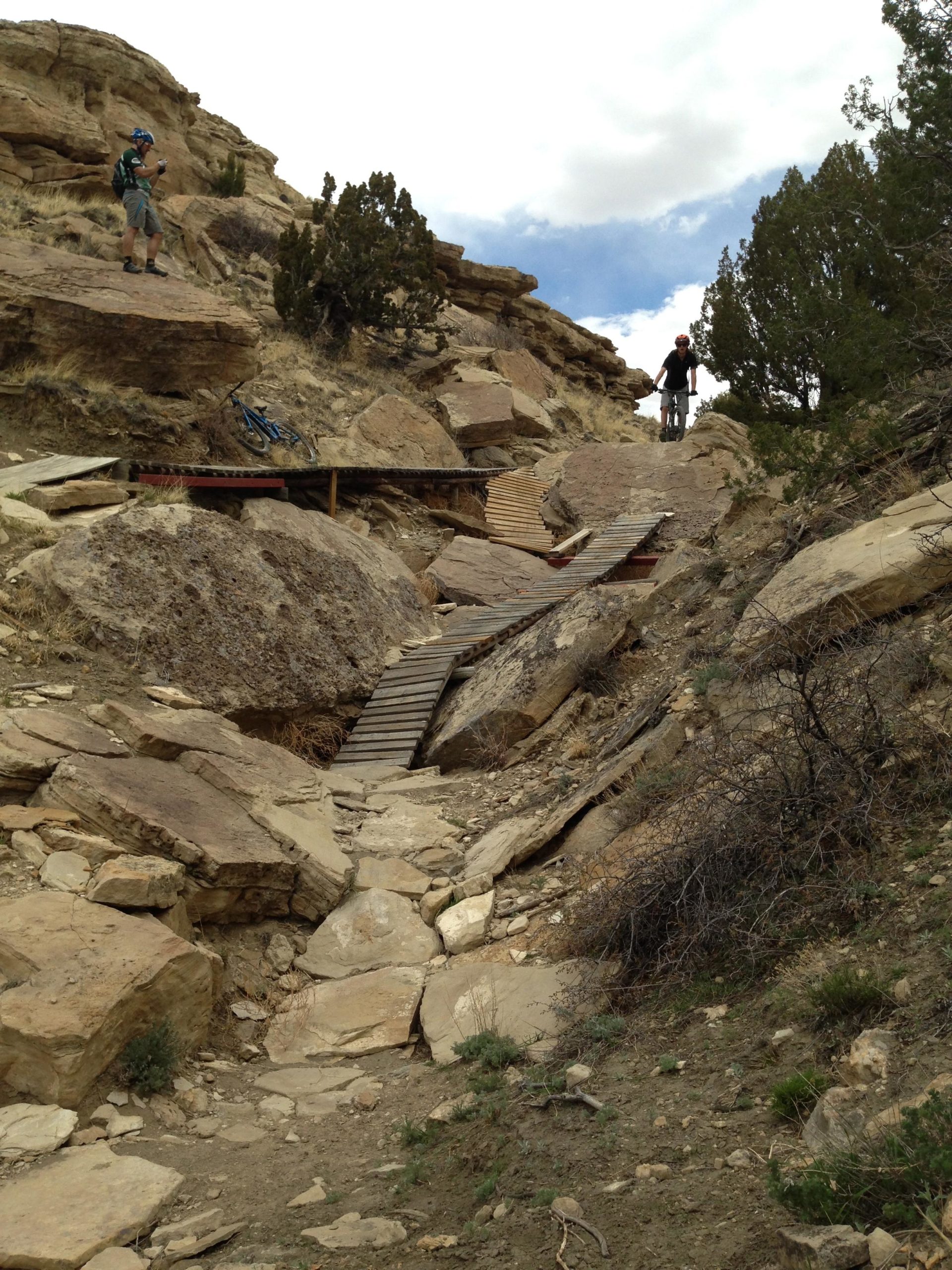 A rugged dirt trail leads through a rocky landscape, with wooden ramps connecting uneven stone pathways. Two mountain bikers are present: one is standing on the hillside, taking a photo, while the other is positioned further up the trail near a rocky outcrop. Sparse vegetation and shrubs dot the terrain beneath a partly cloudy sky. South Shore Lake Pueblo mountain bike trail.