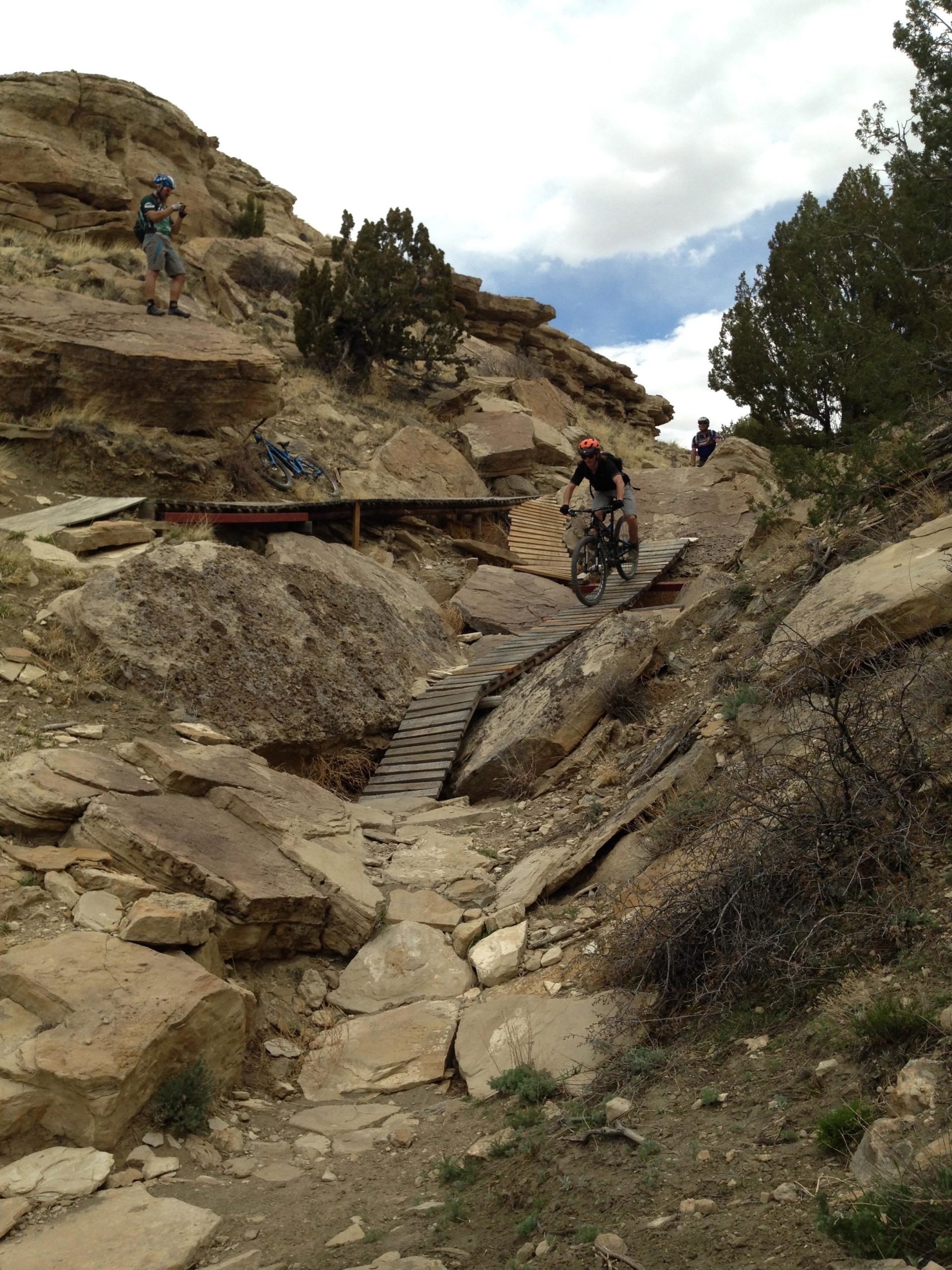 A mountain biker navigates a rocky trail with wooden planks, surrounded by rugged terrain and shrubs, while another rider captures the scene in the background. The sky is partly cloudy, adding to the outdoor atmosphere. South Shore Lake Pueblo mountain bike trail.