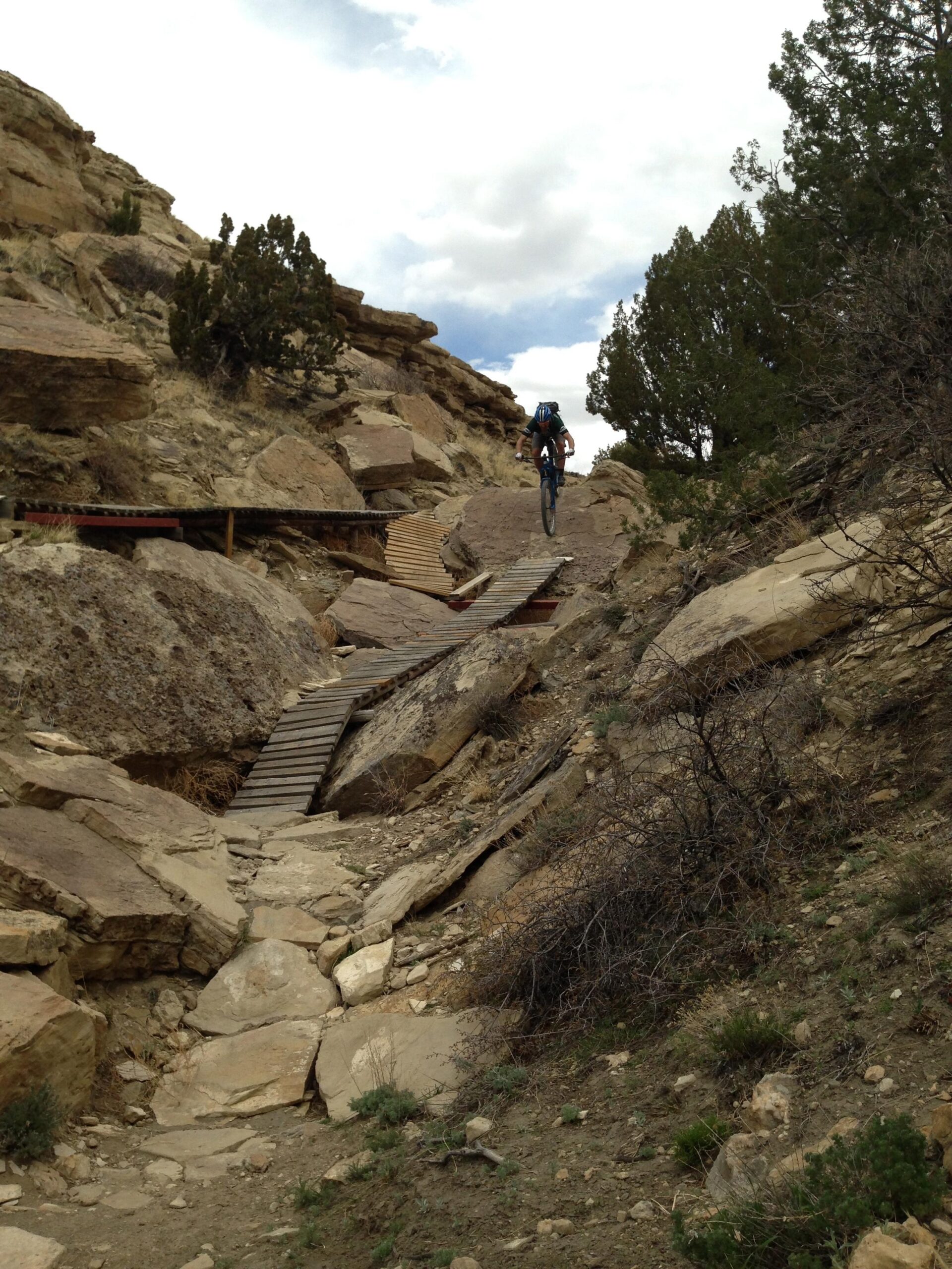 A mountain biker navigates a rugged trail surrounded by rocky terrain and sparse vegetation. A narrow wooden bridge spans a gap in the rocks, leading the cyclist further along the path. The sky is partly cloudy, with patches of blue peeking through the clouds. South Shore Lake Pueblo mountain bike trail.