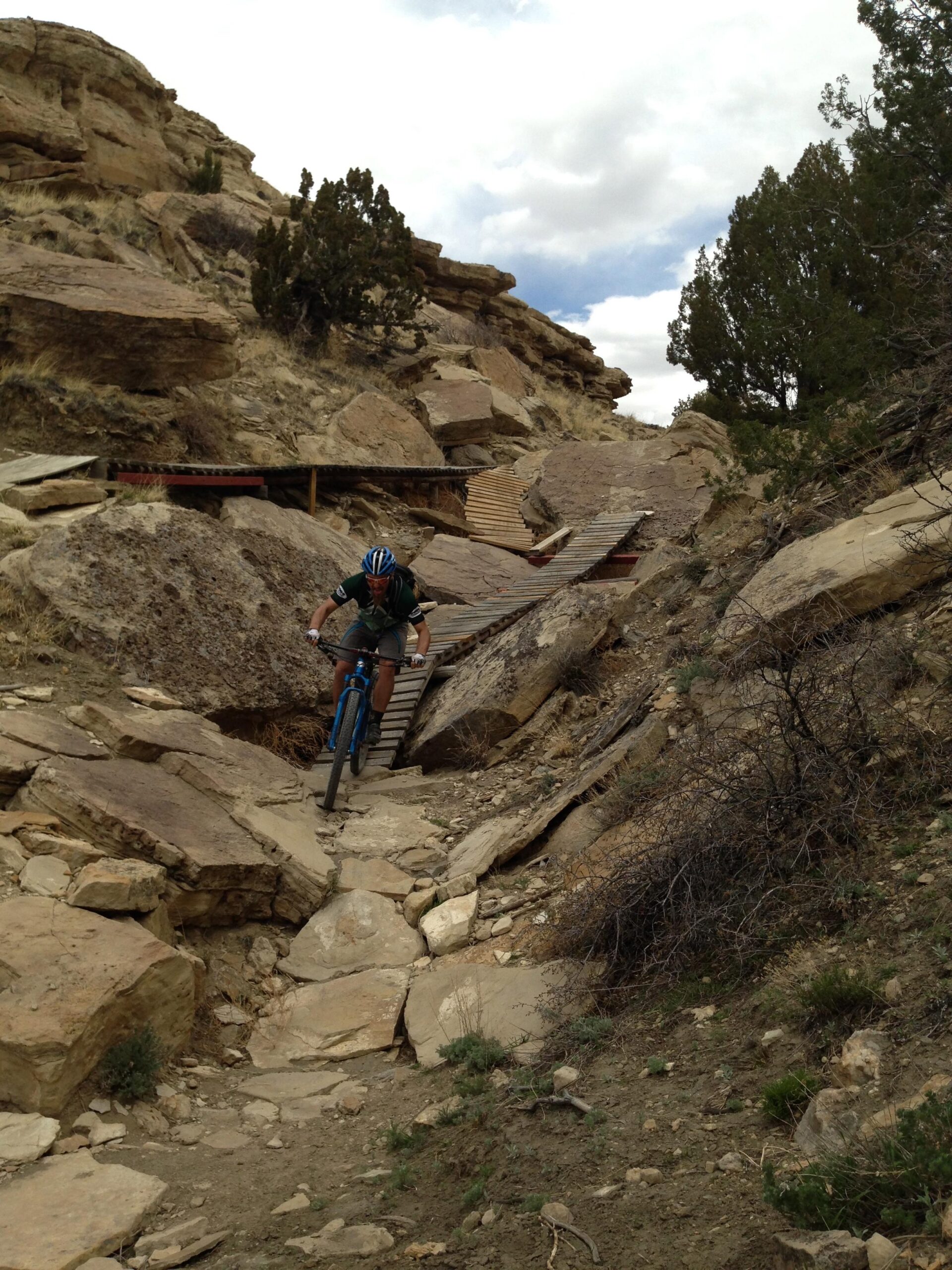A cyclist navigates a rocky mountain biking trail, riding over wooden planks and surrounded by rugged terrain and rocky outcrops under a partly cloudy sky. South Shore Lake Pueblo mountain bike trail.