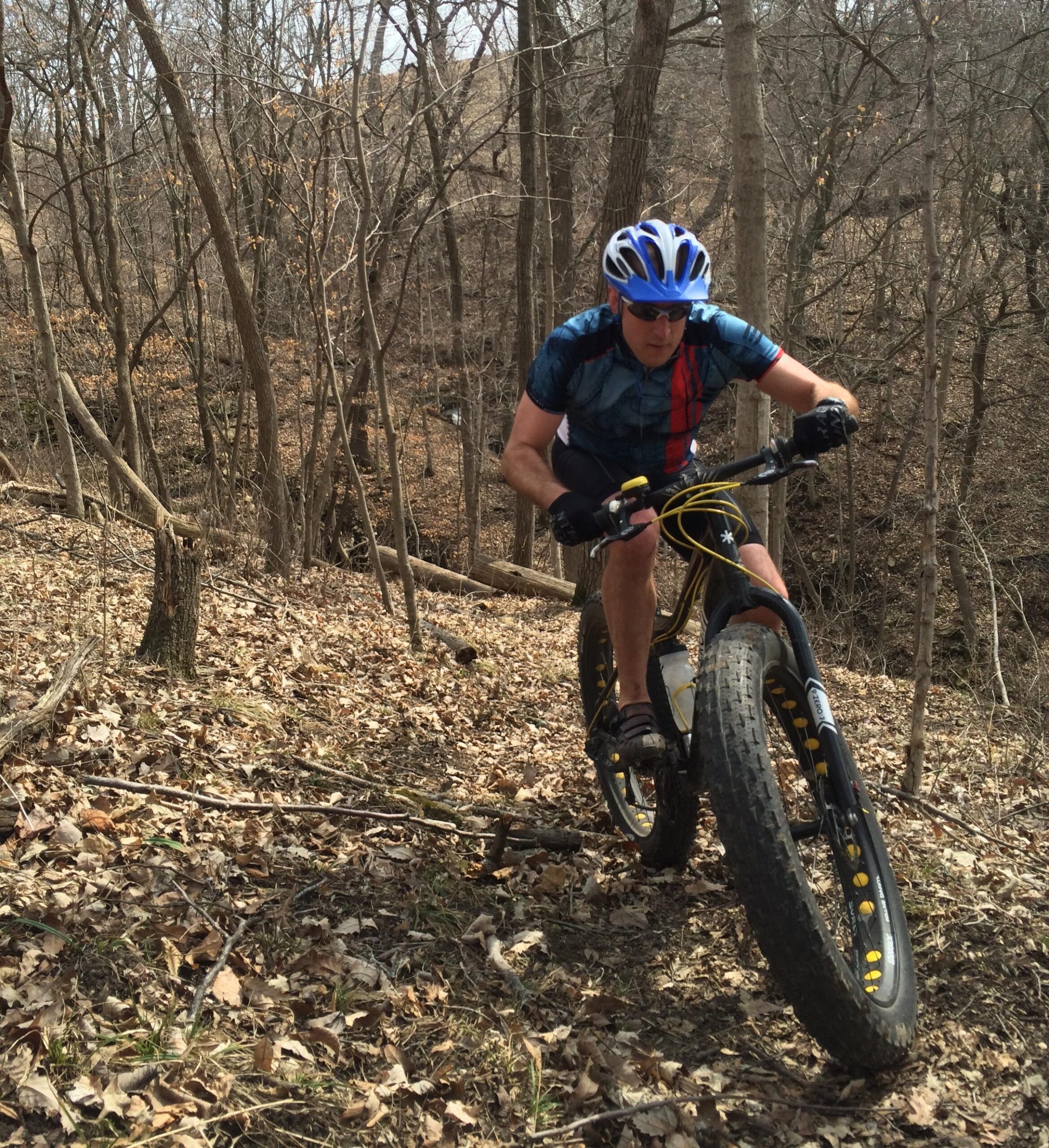 A cyclist navigating a steep, leaf-covered trail in a wooded area. The rider is wearing a blue helmet and sunglasses, riding a fat bike with wide tires, focused on the path ahead. The trees are bare, indicating an early spring or late fall setting.