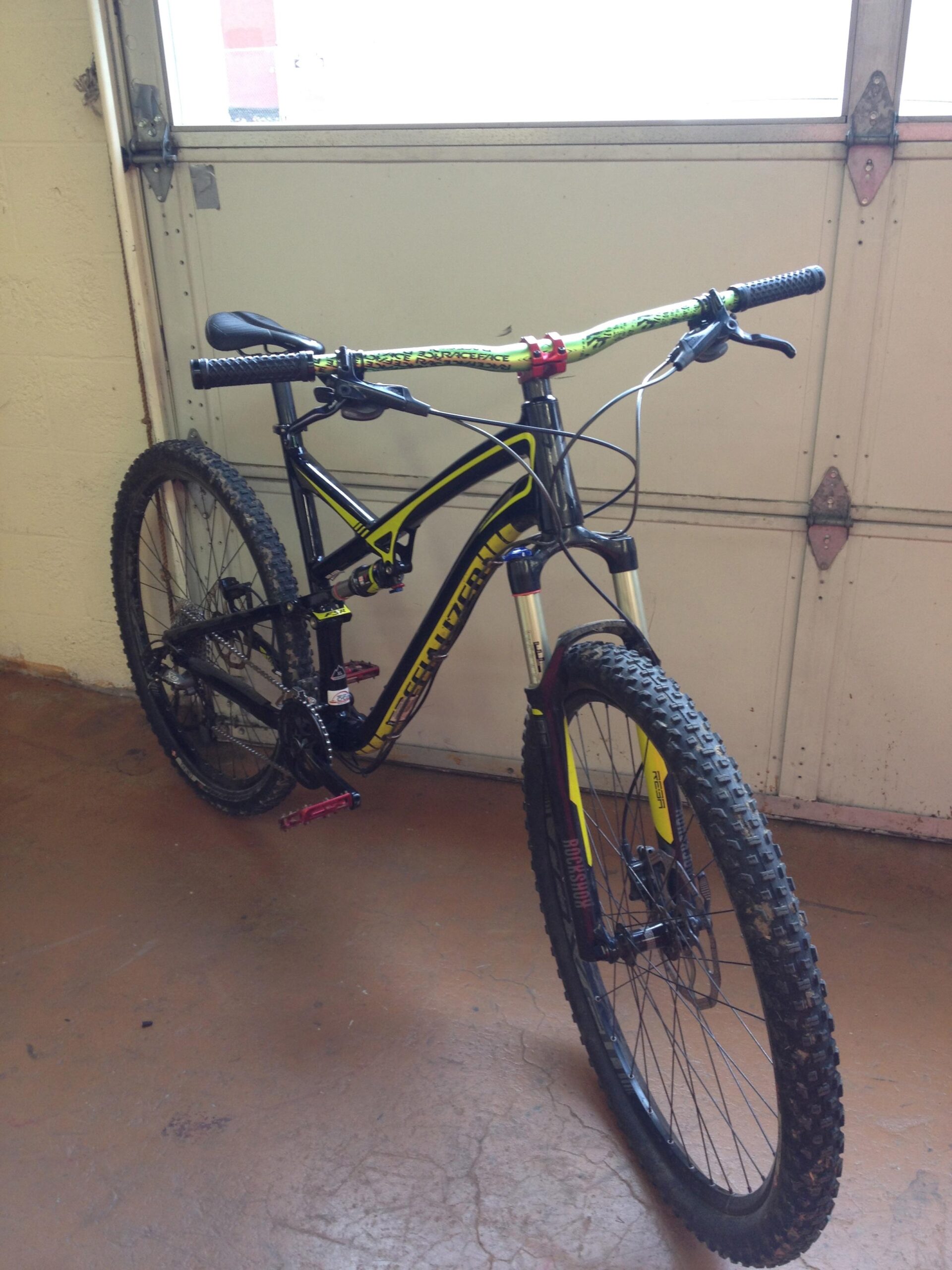 Specialized Camber: A black and yellow mountain bike leaning against a wall in a garage. The bike features wide tires with substantial tread, colorful handlebars, and a visible suspension system. In the background, a partially open garage door allows natural light to enter the space.