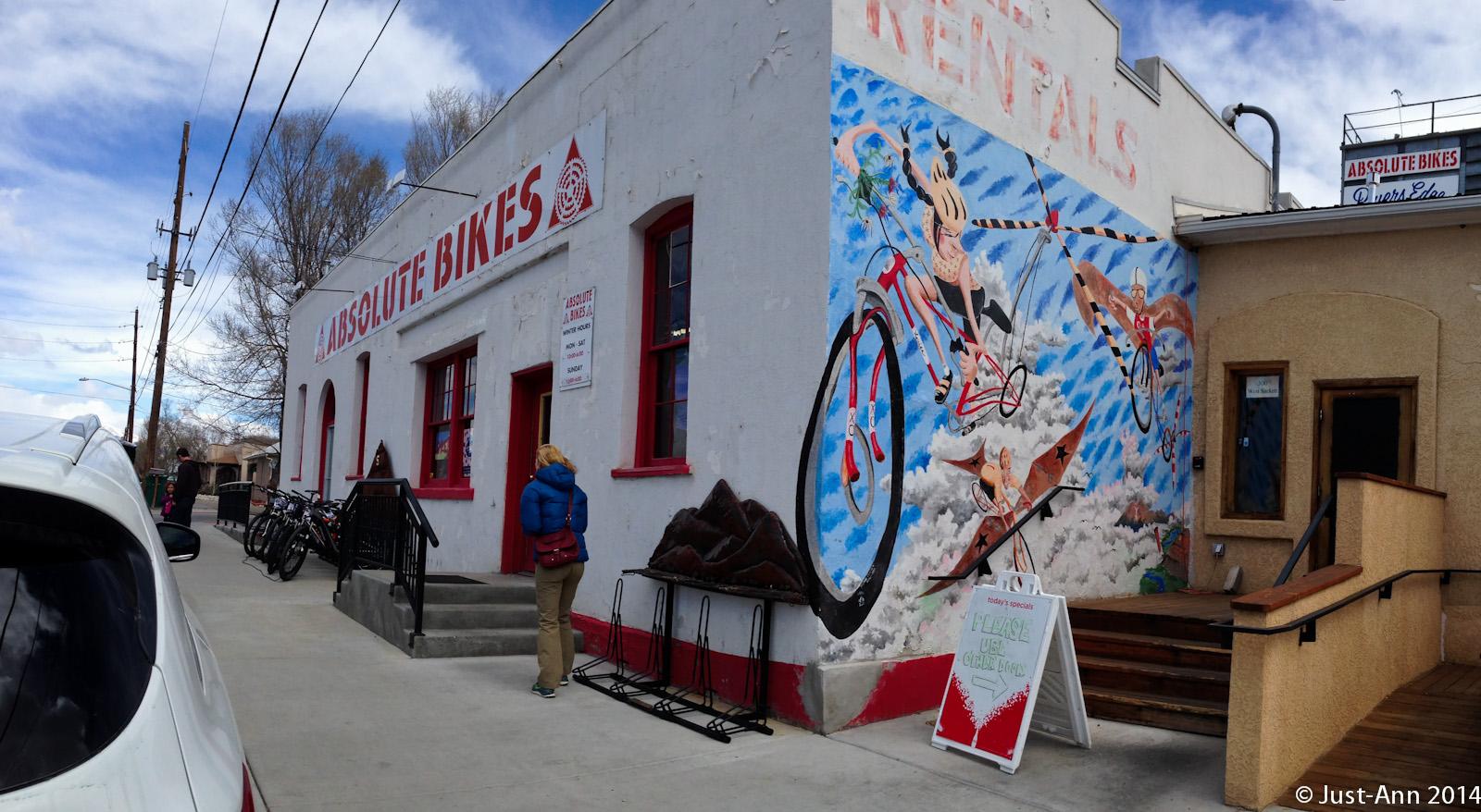 Alt text: "Exterior of Absolute Bikes rental shop, featuring a colorful mural of cyclists against a blue sky. A person stands near the entrance, and bicycles are lined up against the building. The shop has red-framed windows and a sign indicating bike rentals."