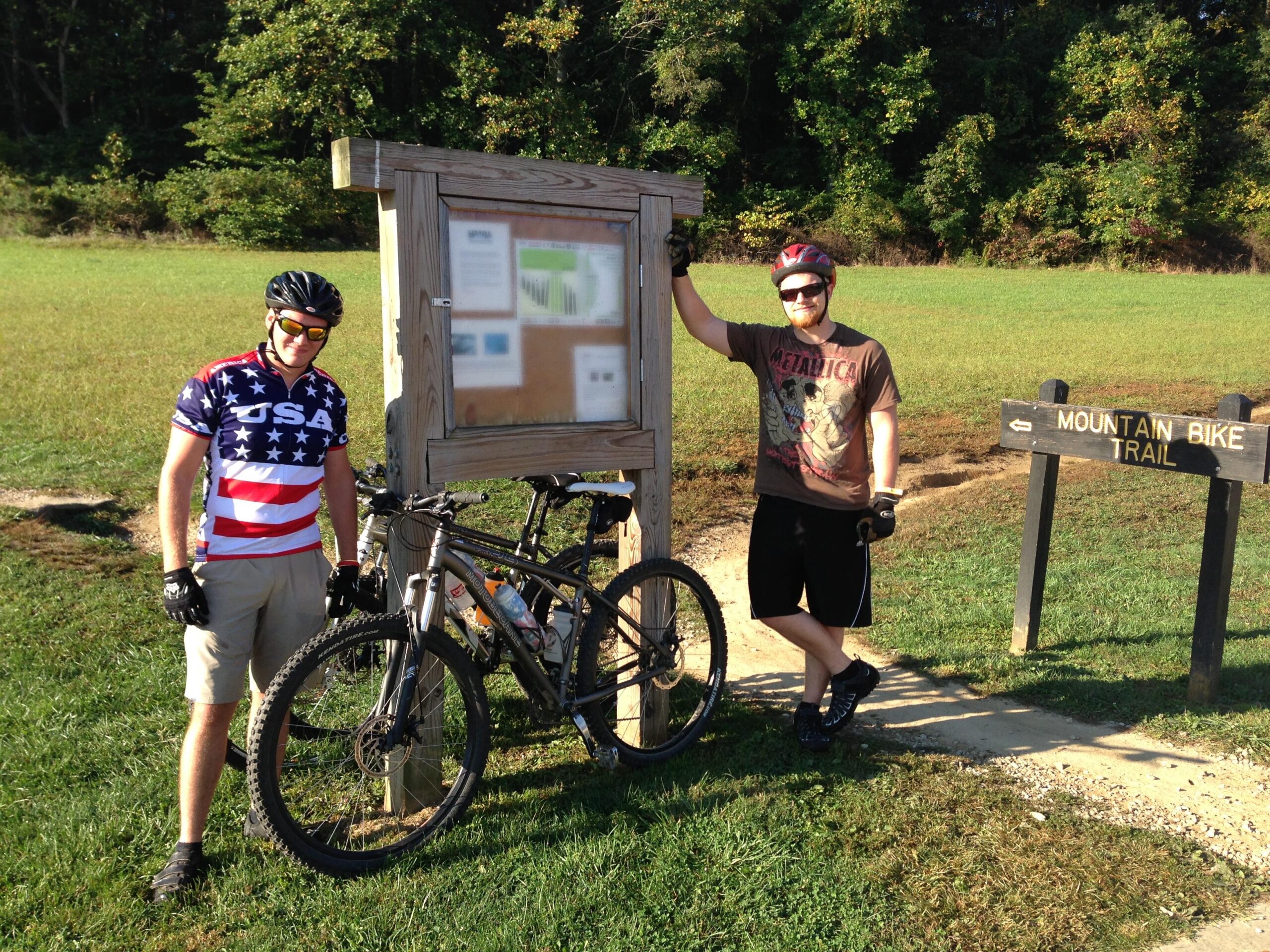 Two mountain bikers pose next to a trail sign labeled "Mountain Bike Trail." One cyclist wears a patriotic "USA" jersey and sunglasses, standing beside a black mountain bike. The other biker, in a Metallica t-shirt, leans against a trail map board. The background features grass and trees under bright sunlight. Brown County Park mountain bike trail.