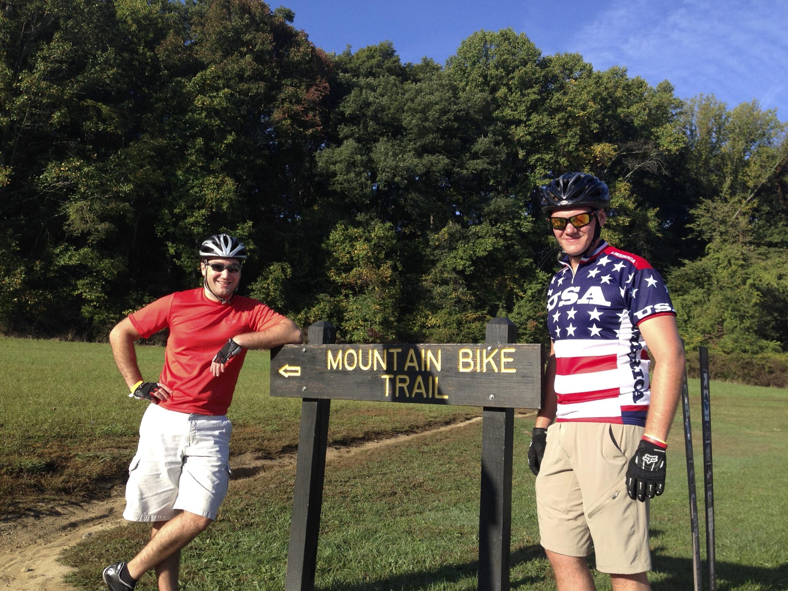 Two men in cycling gear pose next to a sign that reads "Mountain Bike Trail." One man is wearing a red shirt and shorts, while the other is in a patriotic American flag-themed cycling jersey. They are both wearing helmets and sunglasses, standing in a grassy area with trees in the background. The sky is clear and blue. Brown County Park mountain bike trail.