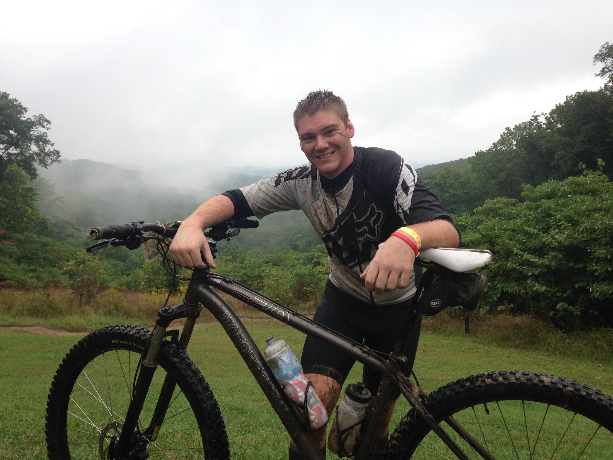 A young man smiles while leaning on his mountain bike in a grassy area surrounded by lush, green hills. He is wearing a muddy, athletic shirt and has dirt smudges on his face, suggesting he has just finished a mountain biking trail. The background features a misty, overcast sky, adding to the outdoor atmosphere. Brown County Park mountain bike trail.