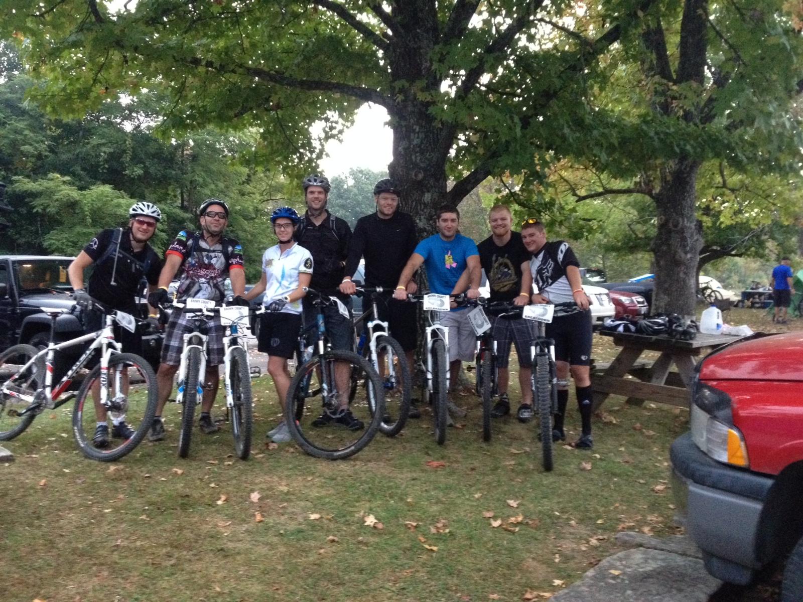 A group of seven mountain bikers, wearing helmets and cycling gear, stand beside their bikes in a park-like setting. They are gathered under a large tree with grassy ground around them. Some vehicles are visible in the background, and the scene captures a camaraderie among the cyclists, likely before a ride or event. Brown County Park mountain bike trail.