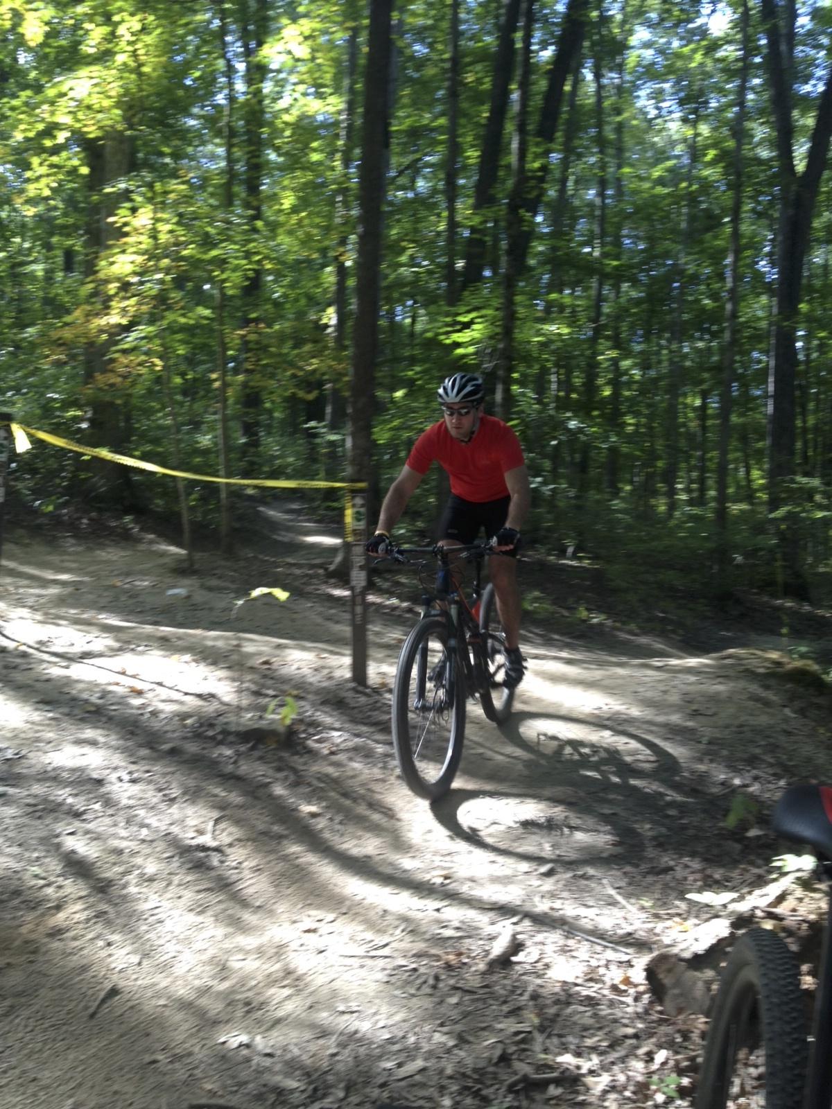 A mountain biker in a red shirt rides along a dirt trail through a wooded area, surrounded by green trees and sunlight filtering through the leaves. Yellow caution tape is visible along the path. Brown County Park mountain bike trail.