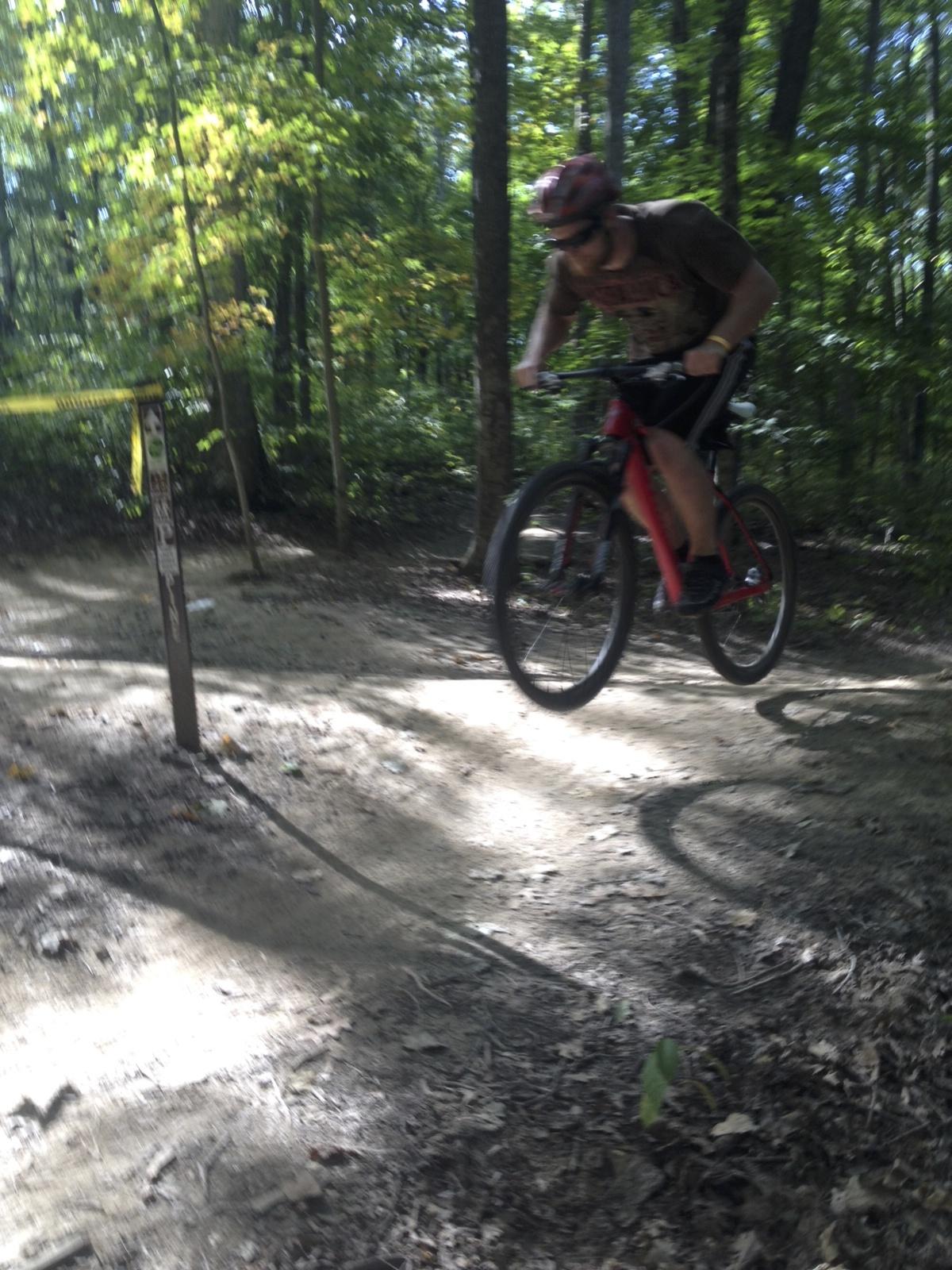 A mountain biker performing a jump on a dirt trail in a wooded area, with trees and sunlight filtering through the foliage. The biker is wearing a helmet and riding a red bike, with a caution tape marker visible in the background. Brown County Park mountain bike trail.