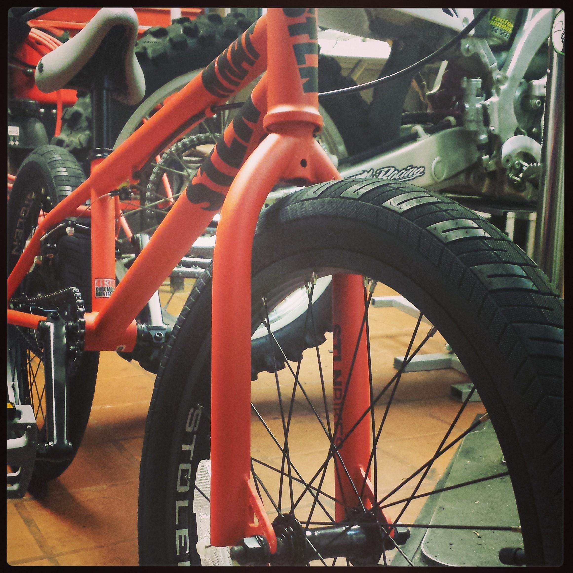 Close-up view of a bright orange bicycle frame with a focus on the front tire and fork, showcasing the bike's design details. The background includes tools and other bike parts, suggesting a workshop setting.