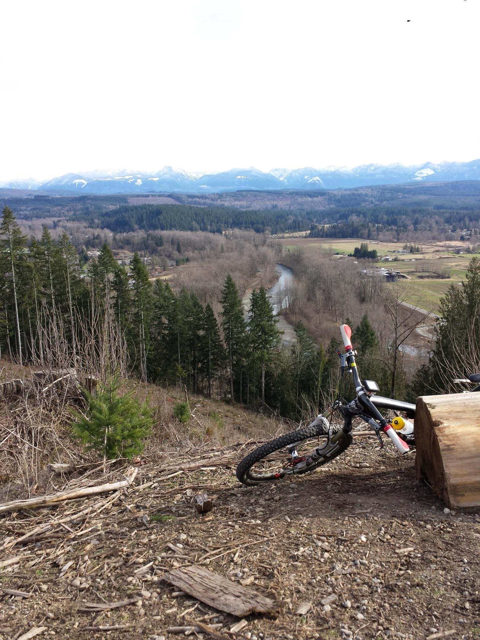 A mountain bike leaning against a log on a hillside, overlooking a scenic valley with a river winding through lush green fields and forests, framed by distant snow-capped mountains under a cloudy sky. Tolt MacDonald mountain bike trail.