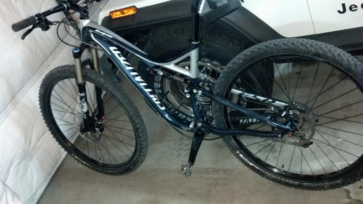Specialized Stumpjumper FSR Comp 29: A mountain bike with a black and silver frame is positioned in a garage. The bike features thick tires and front suspension, with visible dirt on the wheels, indicating recent use. In the background, part of a white vehicle is visible. The setting is indoors with a neutral-colored wall.