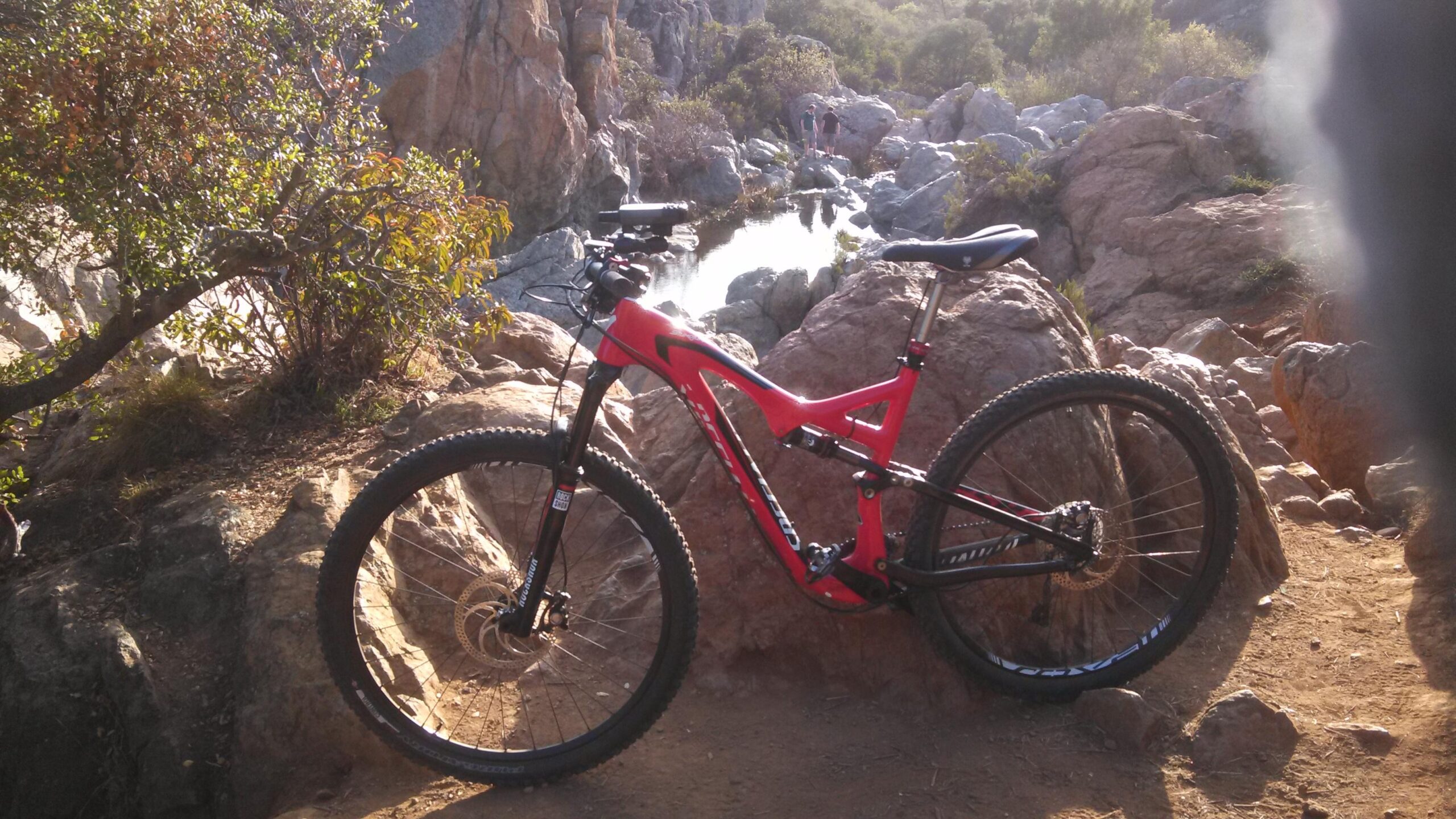 Specialized Stumpjumper FSR Comp Carbon 29: A vibrant red mountain bike resting on rocky terrain with a small waterfall and lush greenery in the background. The scene captures a rugged outdoor setting, ideal for biking enthusiasts.