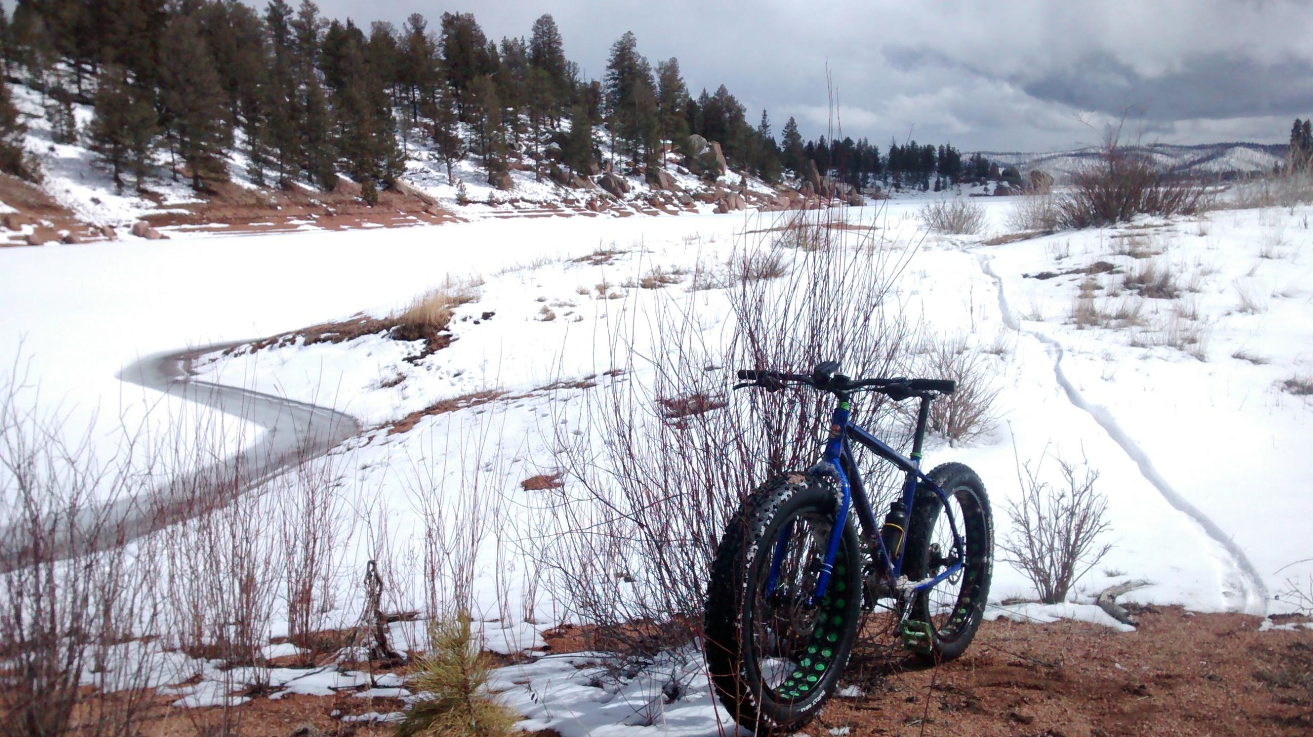 A blue fat bike is parked on a snowy shore beside a frozen lake, surrounded by sparse vegetation and evergreen trees in the background. The scene is set under a cloudy sky, indicating a chilly winter day. A winding path is visible through the snow, leading into the distance. Rampart Reservoir mountain bike trail.