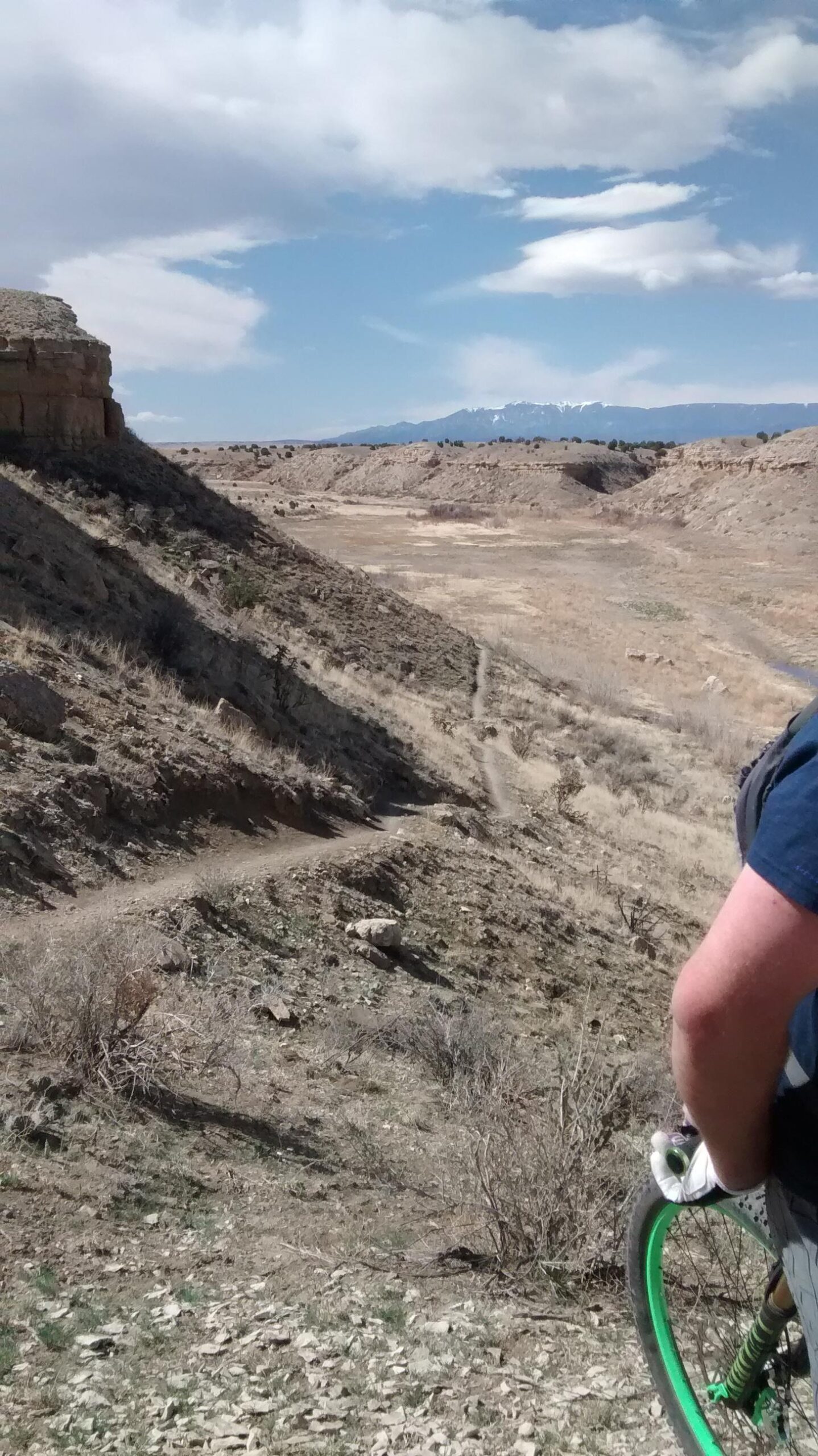 A mountain biker standing near a rugged, dry landscape with hills and a distant mountain range under a partly cloudy sky. A narrow dirt trail winds through the scenery. The biker is holding a bicycle with green wheels, and the foreground features rocky terrain and sparse vegetation. South Shore Lake Pueblo mountain bike trail.
