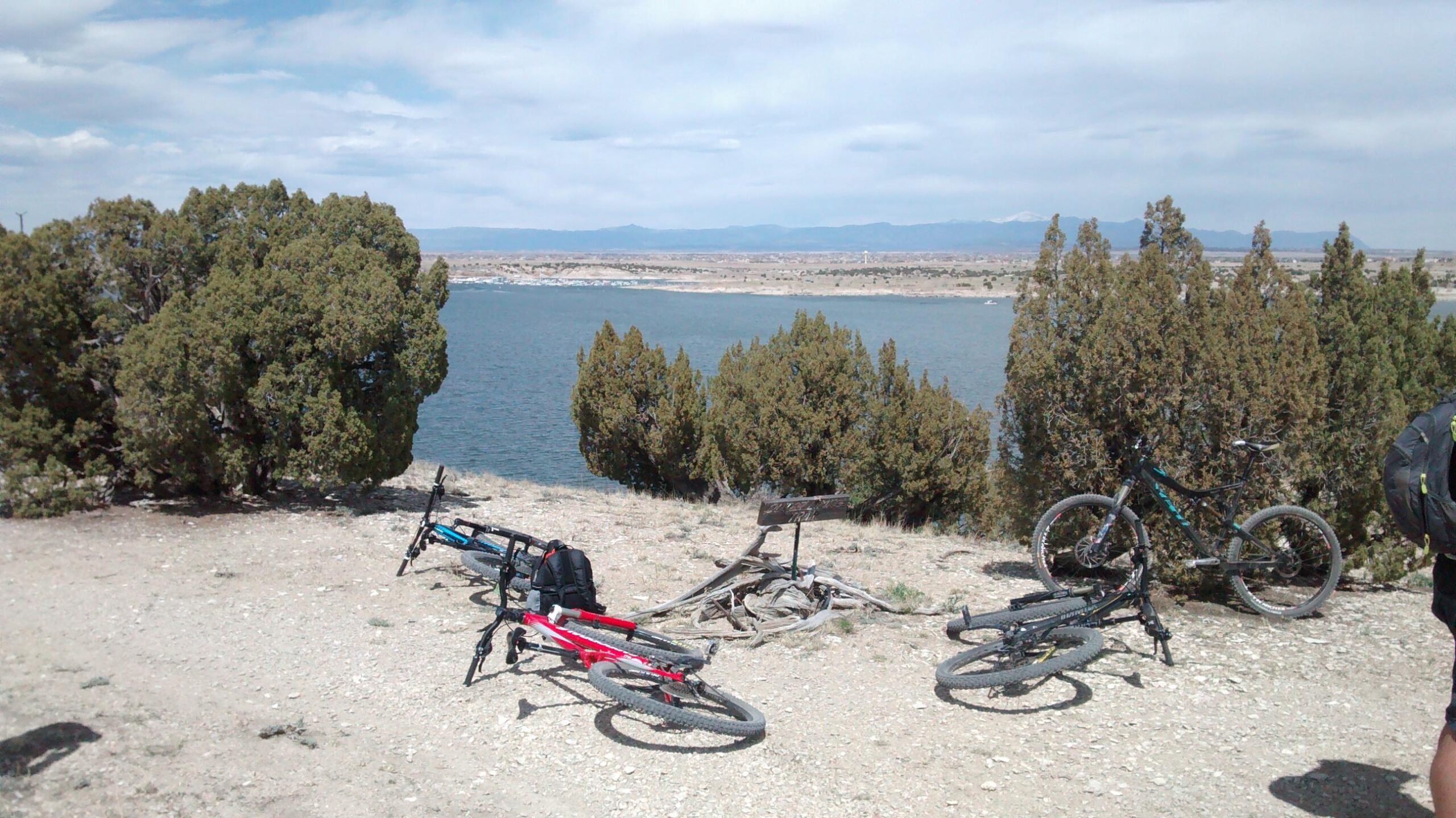 Two mountain bikes are lying on the ground near a scenic lakeside viewpoint surrounded by shrubs. The lake reflects a cloudy sky, and distant mountains are visible in the background, suggesting a peaceful outdoor setting ideal for biking and nature appreciation. South Shore Lake Pueblo mountain bike trail.