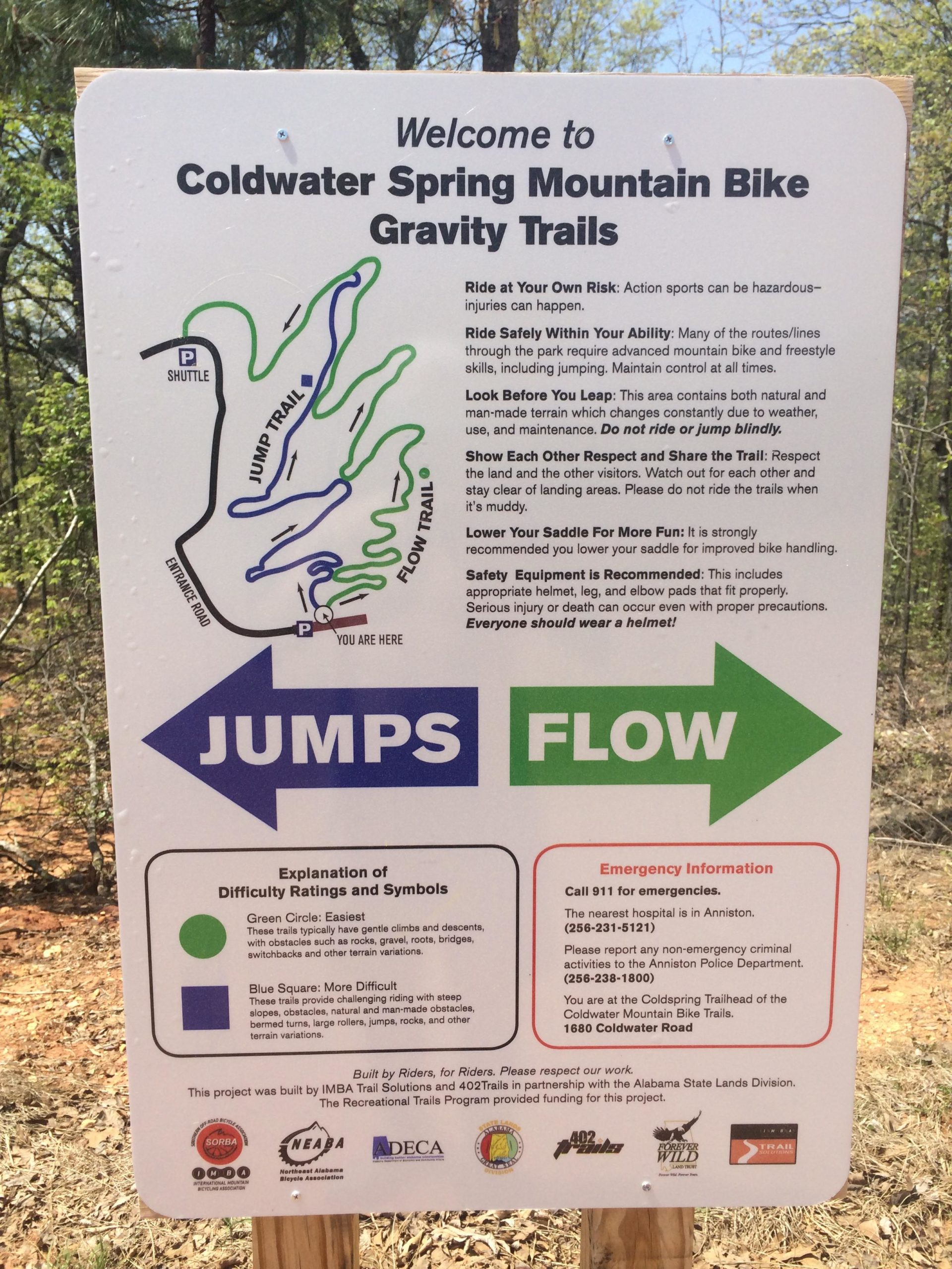 A sign at Coldwater Spring Mountain Bike Gravity Trails providing information about the trails, including safety guidelines, difficulty ratings, and emergency contacts. The map shows the layout of trails labeled "Jump Trail" and "Flow Trail" with color-coded difficulty levels, alongside important reminders for riders about risk and equipment. Coldwater Mountain mountain bike trail.