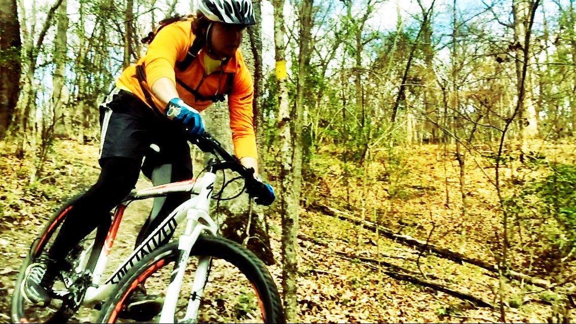A mountain biker in an orange jacket and gloves rides along a trail through a wooded area with fallen leaves and trees in the background. The rider is focused and positioned in a dynamic pose, showcasing the thrill of mountain biking. Wildwood Trails mountain bike trail.