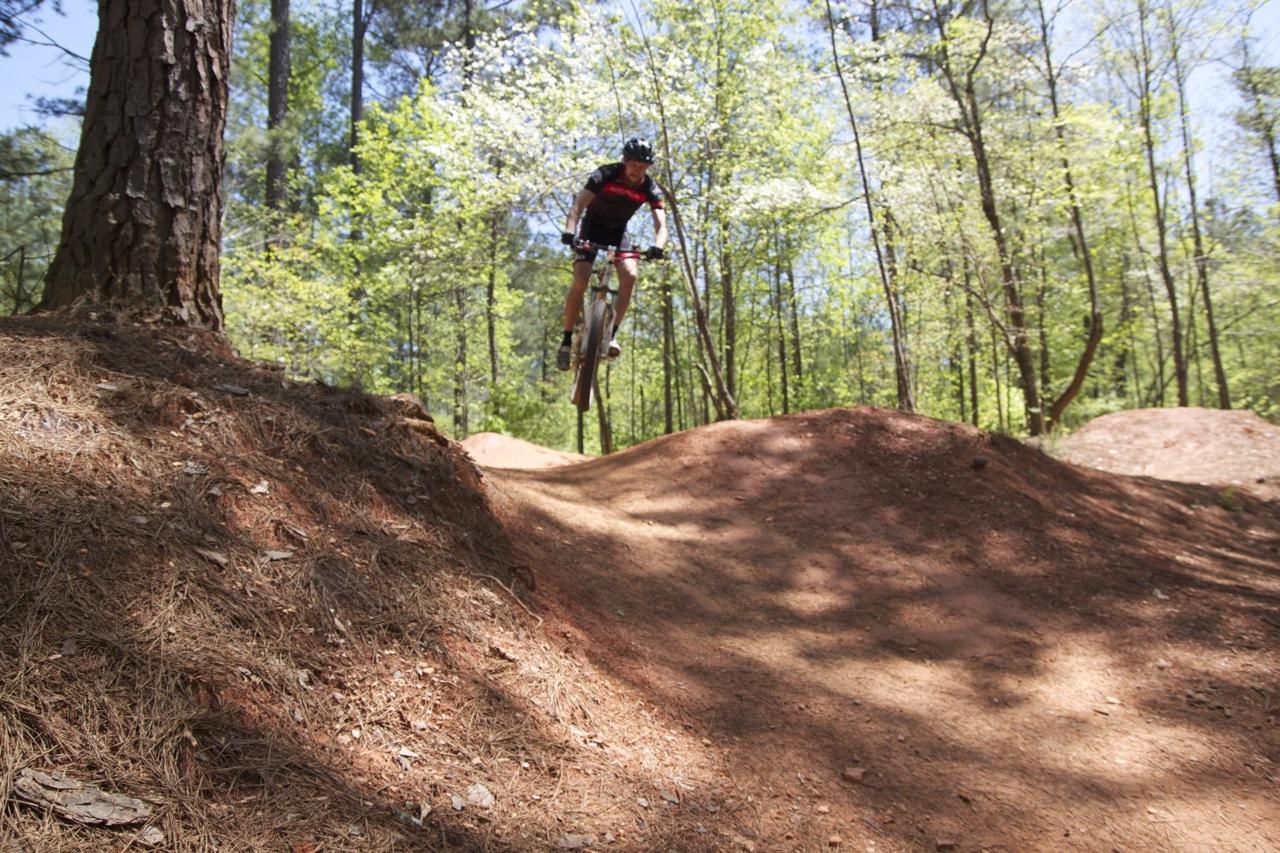 A cyclist mid-jump over a dirt ramp in a wooded area, surrounded by green trees and sunlight filtering through the leaves. The terrain consists of well-formed dirt mounds, indicating a mountain biking course. Blankets Creek mountain bike trail.