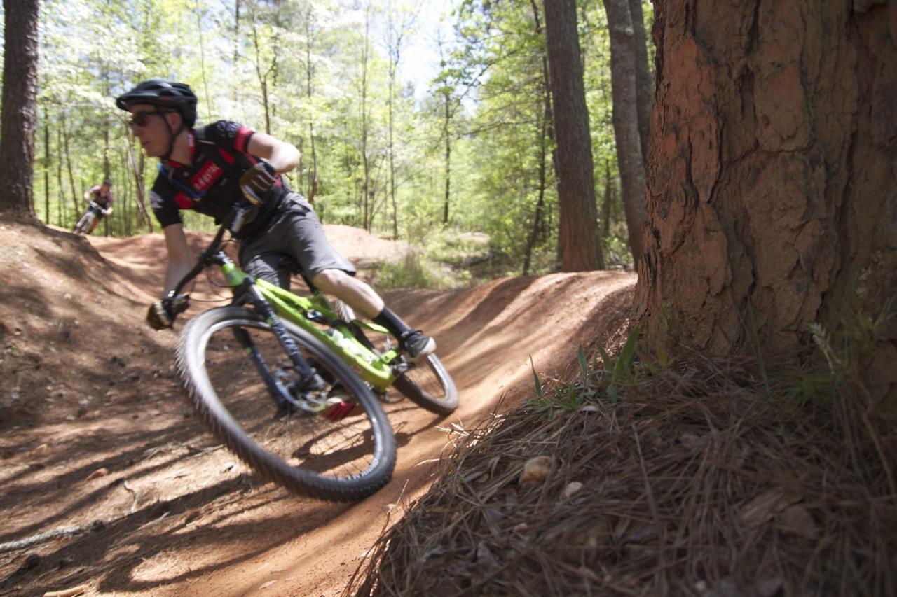 A cyclist riding a green mountain bike through a winding dirt trail in a wooded area, with trees and vibrant greenery surrounding the path. Another cyclist can be seen in the background, also navigating the trail. Quehl Holler mountain bike trail.
