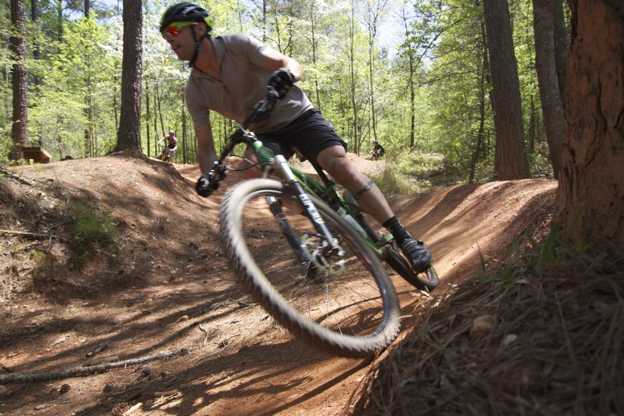 A mountain biker leans into a turn on a dirt trail surrounded by trees, showcasing dynamic movement and skill. The rider is wearing a helmet and sunglasses, dressed in a short-sleeved shirt and shorts. In the background, another cyclist can be seen riding along the trail. Blankets Creek mountain bike trail.