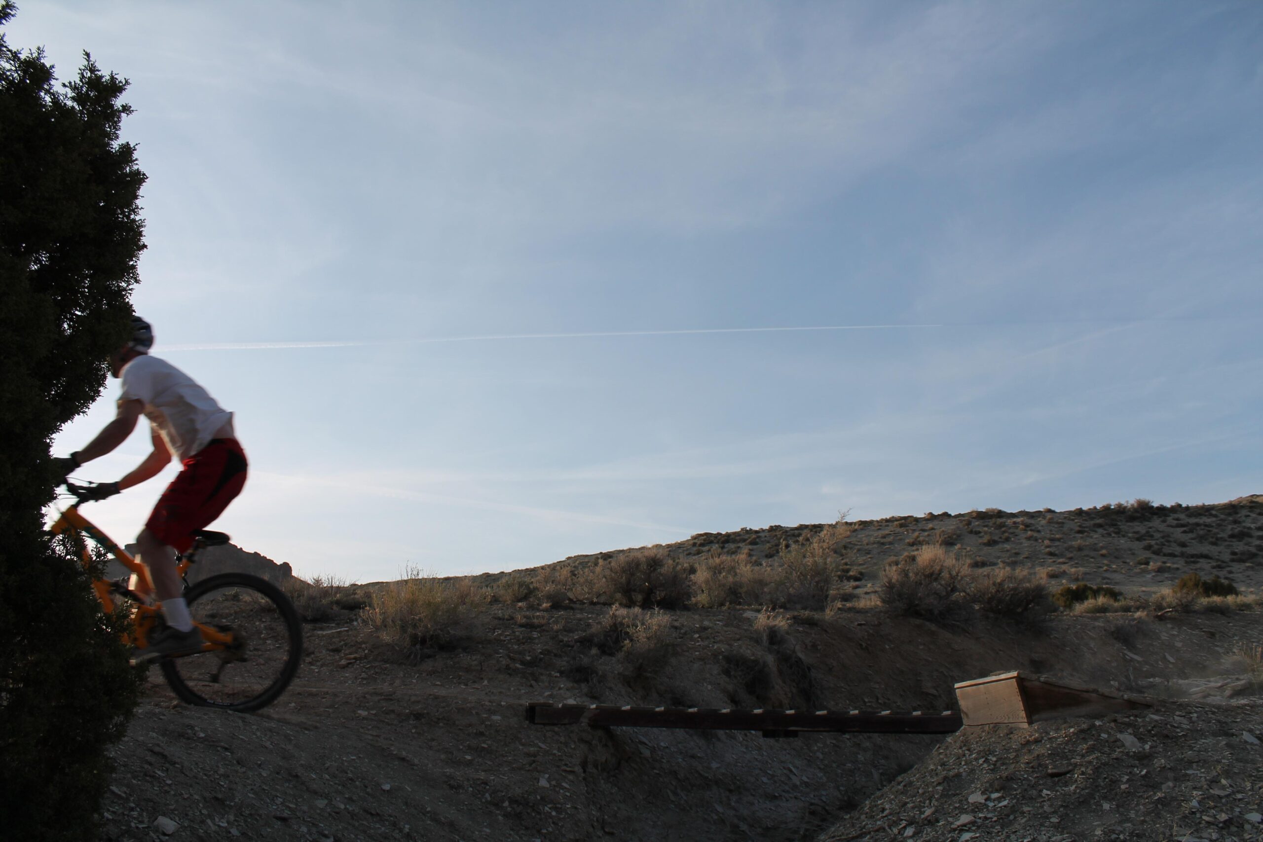 A mountain biker in athletic gear rides over a dirt trail, approaching a small wooden ramp, with a clear blue sky and rocky terrain in the background. Tnt mountain bike trail.