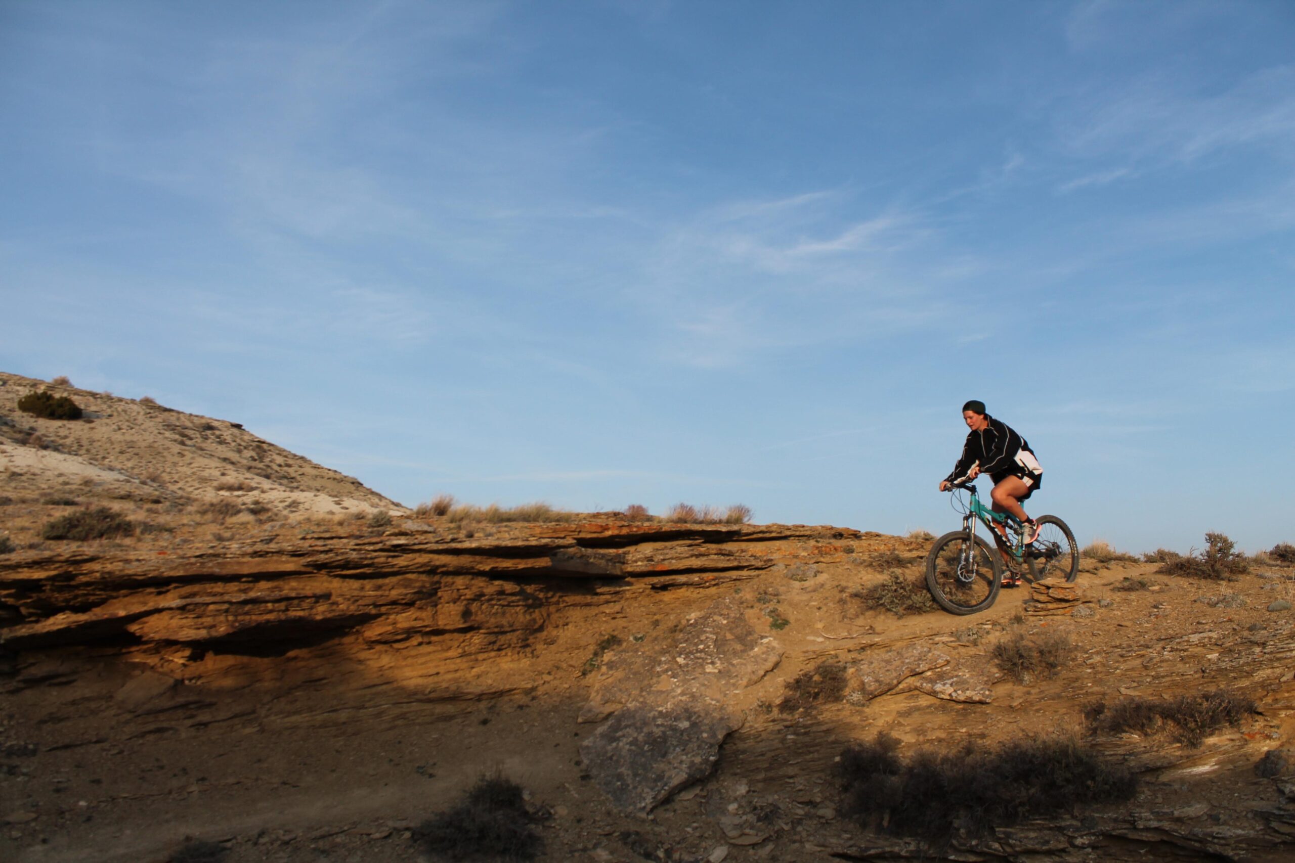 A mountain biker navigating a rocky terrain against a clear blue sky, with a hilly landscape in the background. The rider is wearing a black jacket and shorts, showcasing an adventurous outdoor setting. Brent & Mikes' Trail mountain bike trail.