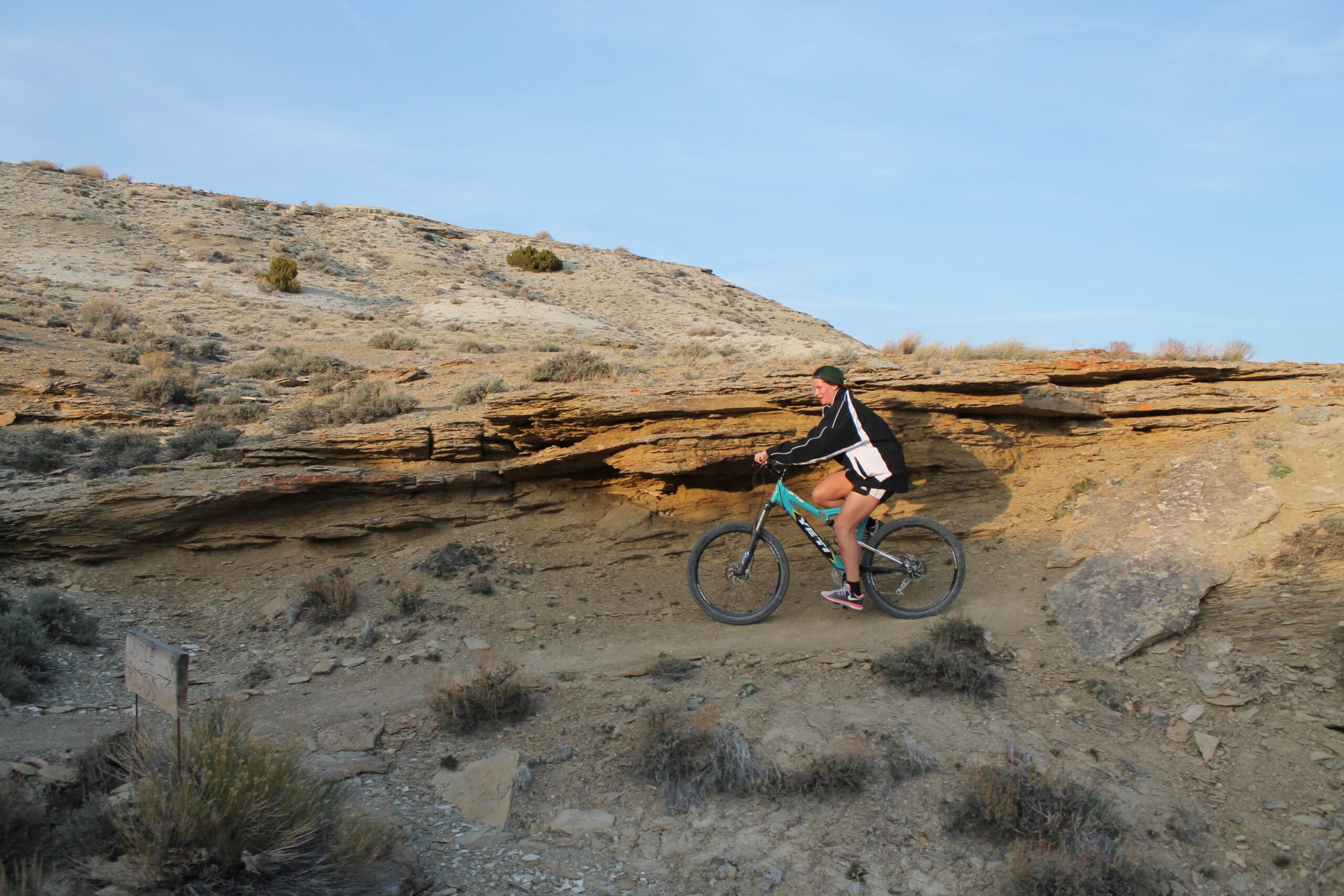 A cyclist riding a mountain bike on a dirt trail surrounded by rocky terrain and sparse vegetation under a blue sky. Brent & Mikes' Trail mountain bike trail.