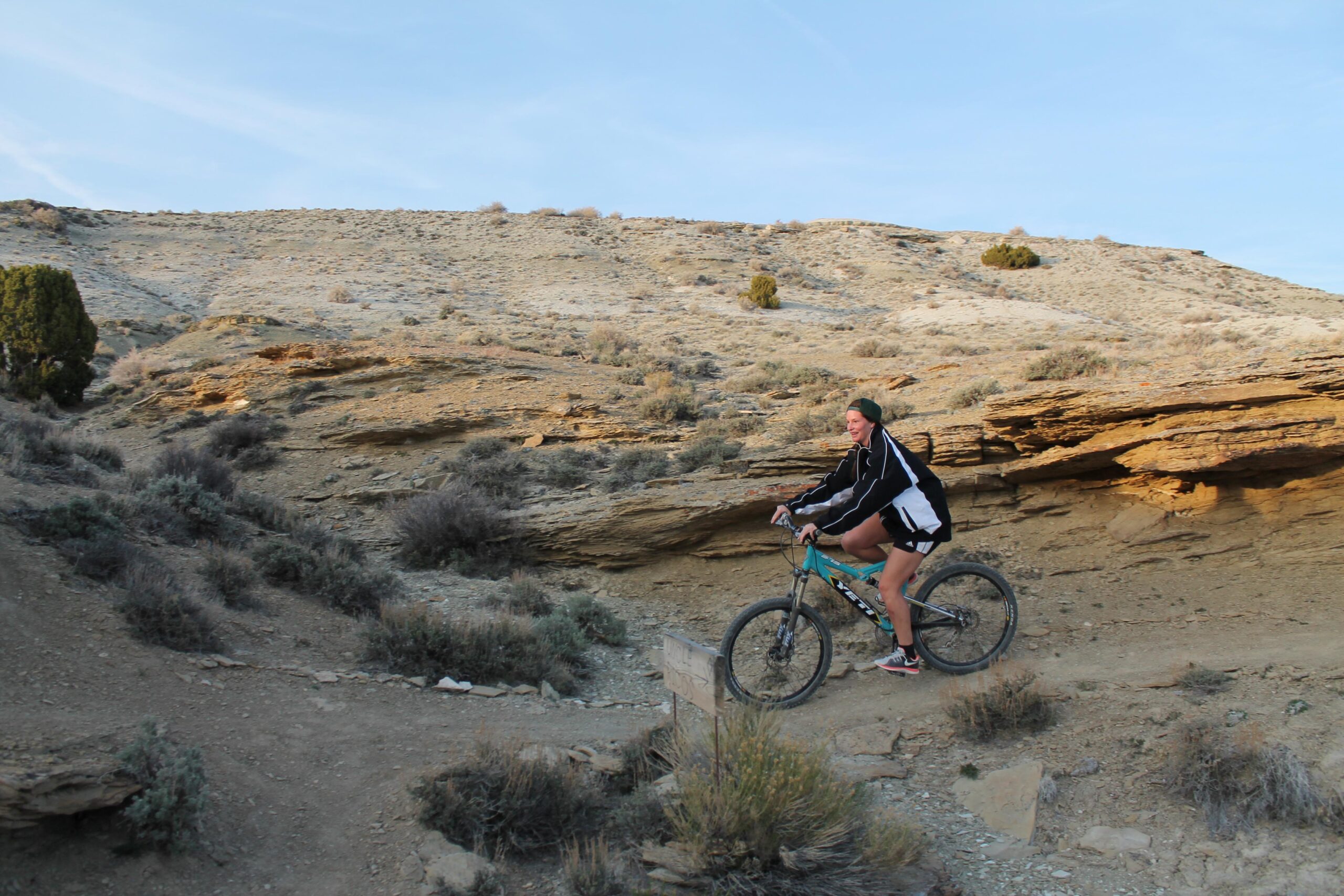A person cycling on a mountain bike along a rocky trail in a desert landscape, with shrubs and uneven terrain. The sun is setting in the background, casting warm light on the hills. Brent & Mikes' Trail mountain bike trail.