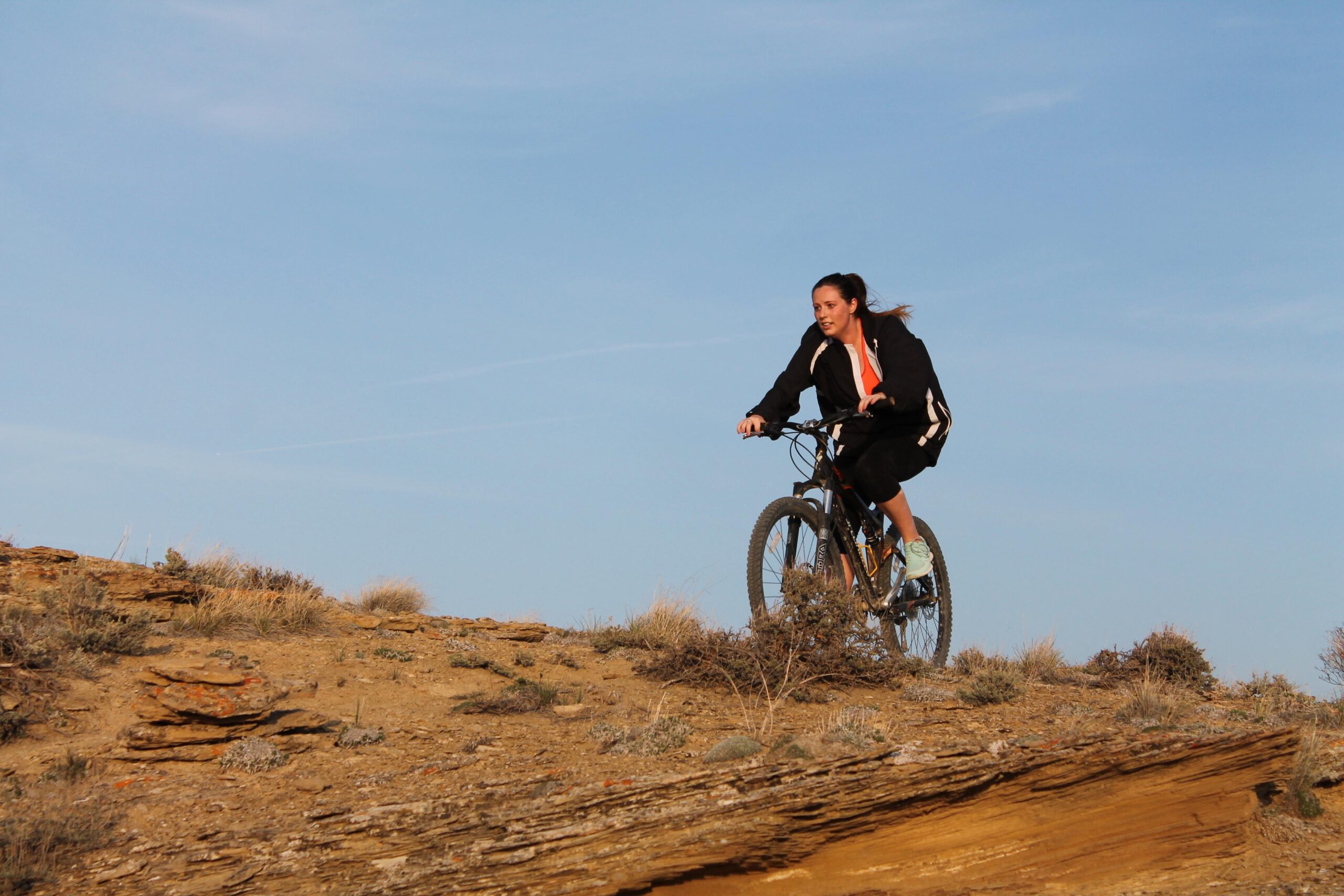 A person riding a mountain bike on a rocky terrain, with dry grass and shrubs in the background under a clear blue sky. The cyclist is wearing a black jacket and orange top, demonstrating skill and agility as they navigate the landscape. Brent & Mikes' Trail mountain bike trail.