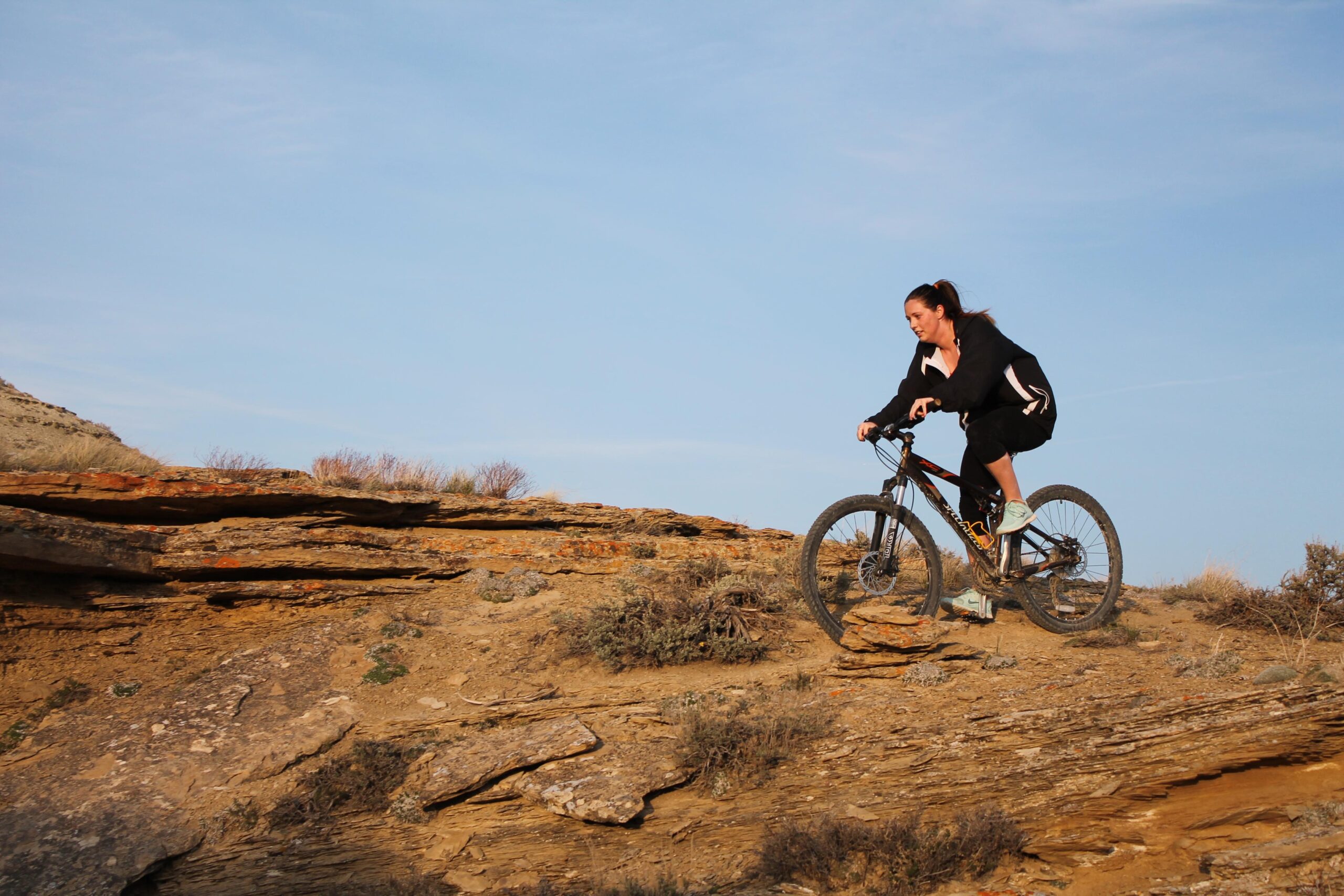 A woman riding a mountain bike on rocky terrain, navigating a slope with shrubs and dry grass in the background, under a blue sky. Brent & Mikes' Trail mountain bike trail.