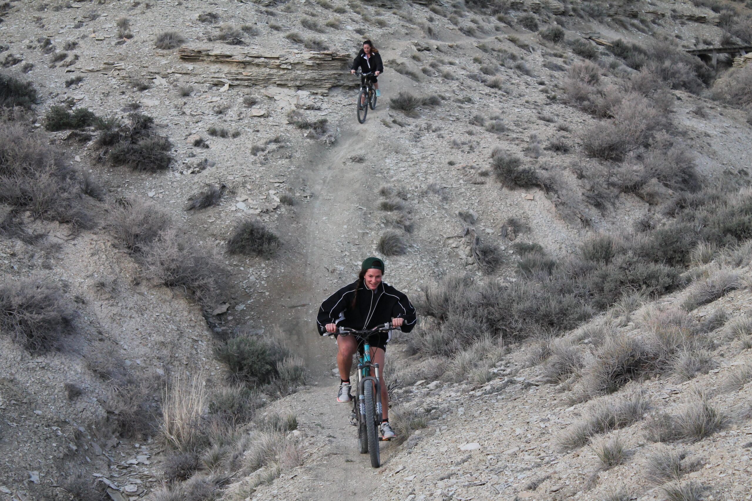 Two mountain bikers riding on a rugged trail in a dry, rocky landscape. One rider is descending from a higher path while the other is climbing up, both surrounded by sparse vegetation. The terrain features rocky outcrops and low shrubs. Uncle Ricos mountain bike trail.