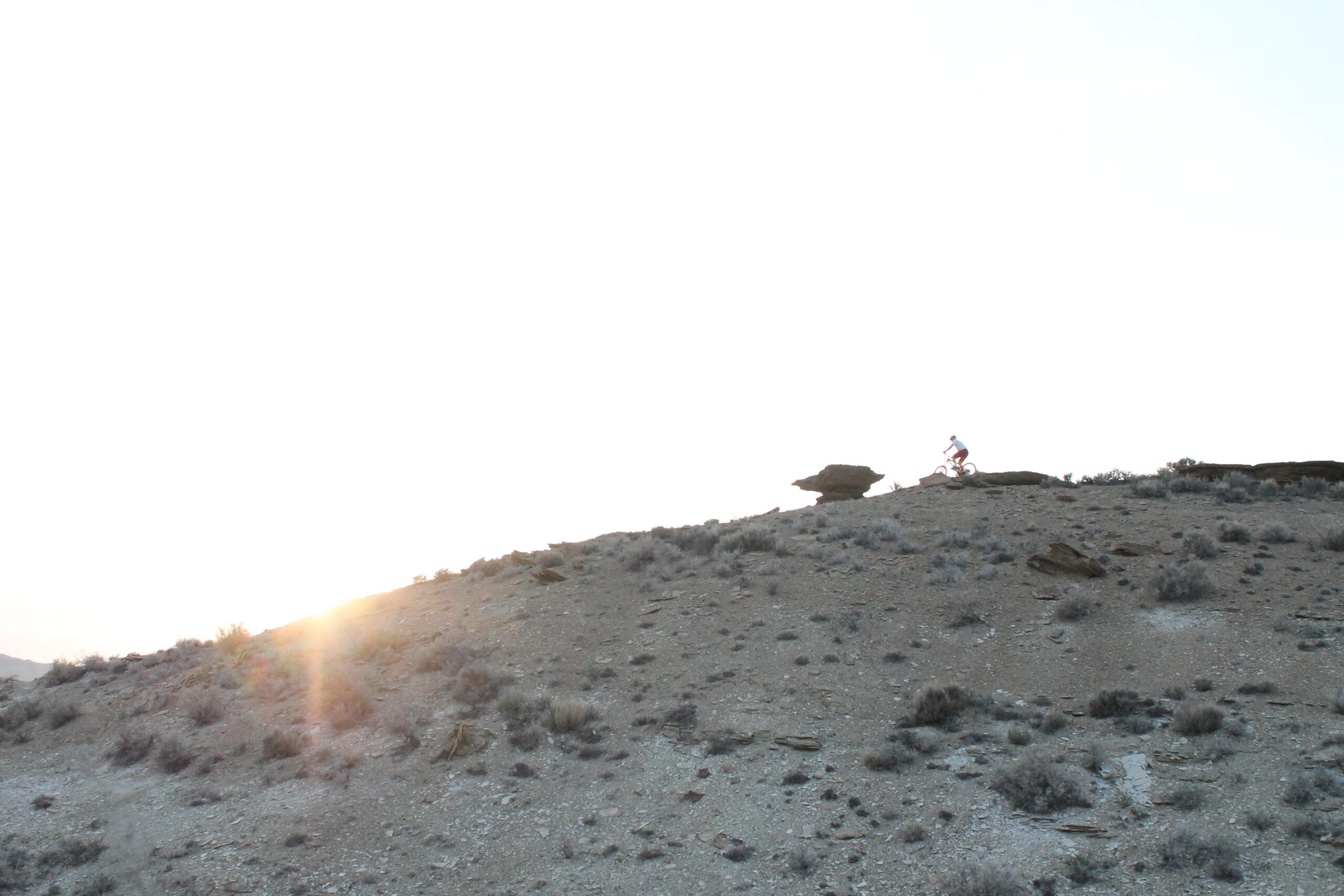 A lone cyclist rides up a dusty hill at sunset, with golden rays of sunlight illuminating the landscape and casting soft shadows across the rocky terrain. Sparse vegetation and small shrubs dot the hillside, creating a serene outdoor scene. Uncle Ricos mountain bike trail.