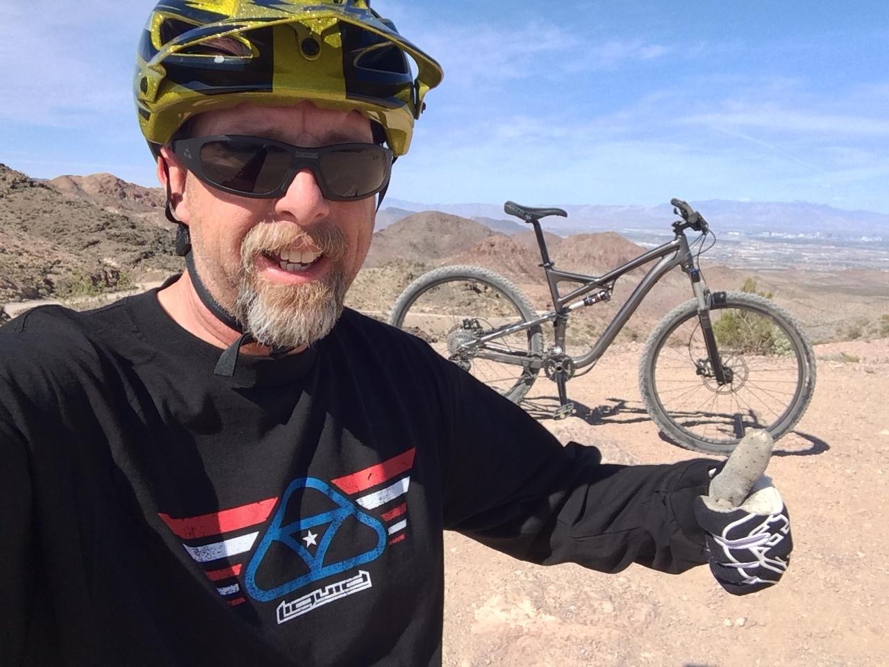 A man wearing a yellow helmet and sunglasses takes a selfie on a mountain biking trail. He is dressed in a black shirt with a colorful logo, holding a rock in one hand while giving a thumbs-up with the other. A mountain bike is parked behind him, with a scenic view of hills and valleys in the background under a clear blue sky. Bootleg Canyon mountain bike trail.