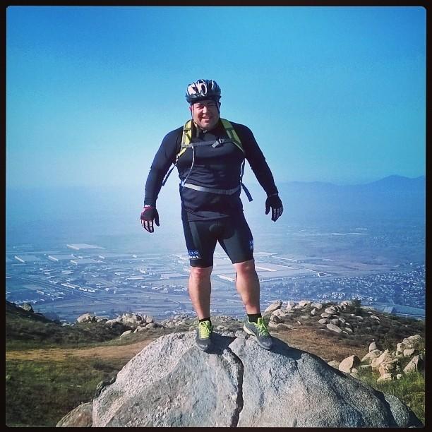 A cyclist standing on a large rock at the summit of a hill, wearing a helmet and athletic gear. The landscape below shows fields and a distant view of the city under a clear blue sky. Box Springs Mountain Access Road mountain bike trail.