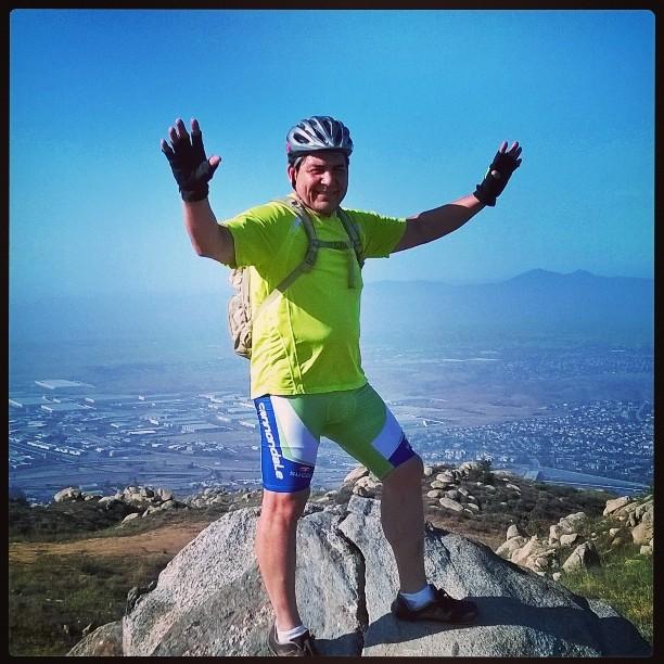 A man in a bright yellow cycling shirt and blue shorts stands triumphantly on a rock at a mountain peak. He is smiling and has his arms raised, clearly celebrating the accomplishment. He is wearing a cycling helmet and has a small backpack. In the background, there is a panoramic view of valleys and distant mountains under a clear blue sky. Box Springs Mountain Access Road mountain bike trail.