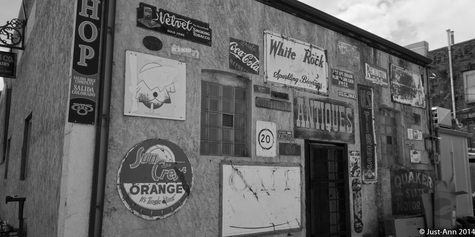A vintage building with a textured wall covered in various old signs and advertisements, displayed in black and white. The signs include brands like Coca-Cola, Sun Orange, and Remington Tires, along with the word "ANTIQUE" prominently featured. Other signs indicate the presence of a bike shop and a café.