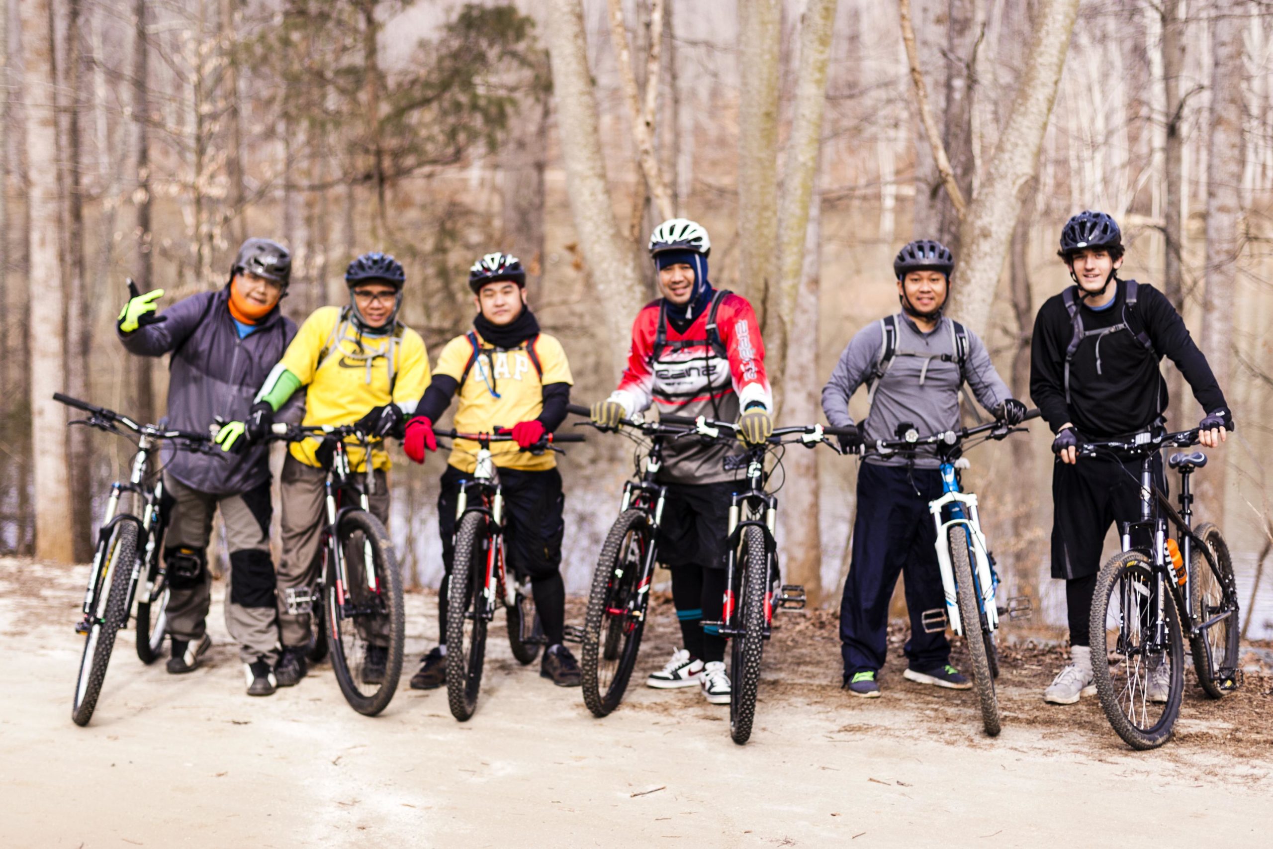 A group of six mountain bikers posing for a photo on a dirt trail surrounded by trees. They are wearing various biking gear, including helmets and gloves, and standing next to their bicycles. The background features a natural setting with a hint of water in the distance. Salem Lake mountain bike trail.
