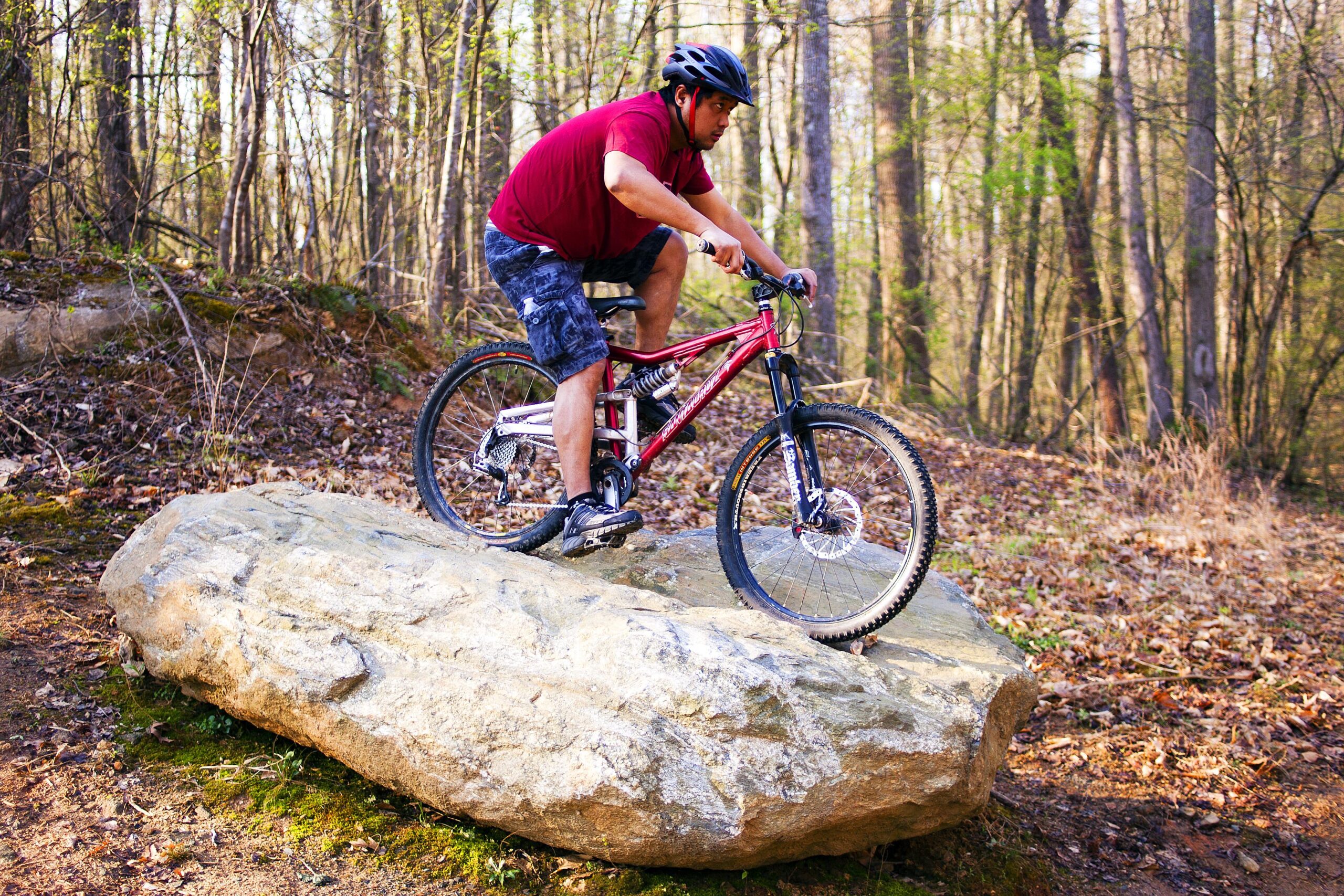 A person riding a mountain bike on top of a large rock in a wooded area, showcasing an adventurous outdoor activity. The individual is wearing a helmet and is positioned in a dynamic stance, suggesting skill and control while navigating the terrain. Salem Lake mountain bike trail.
