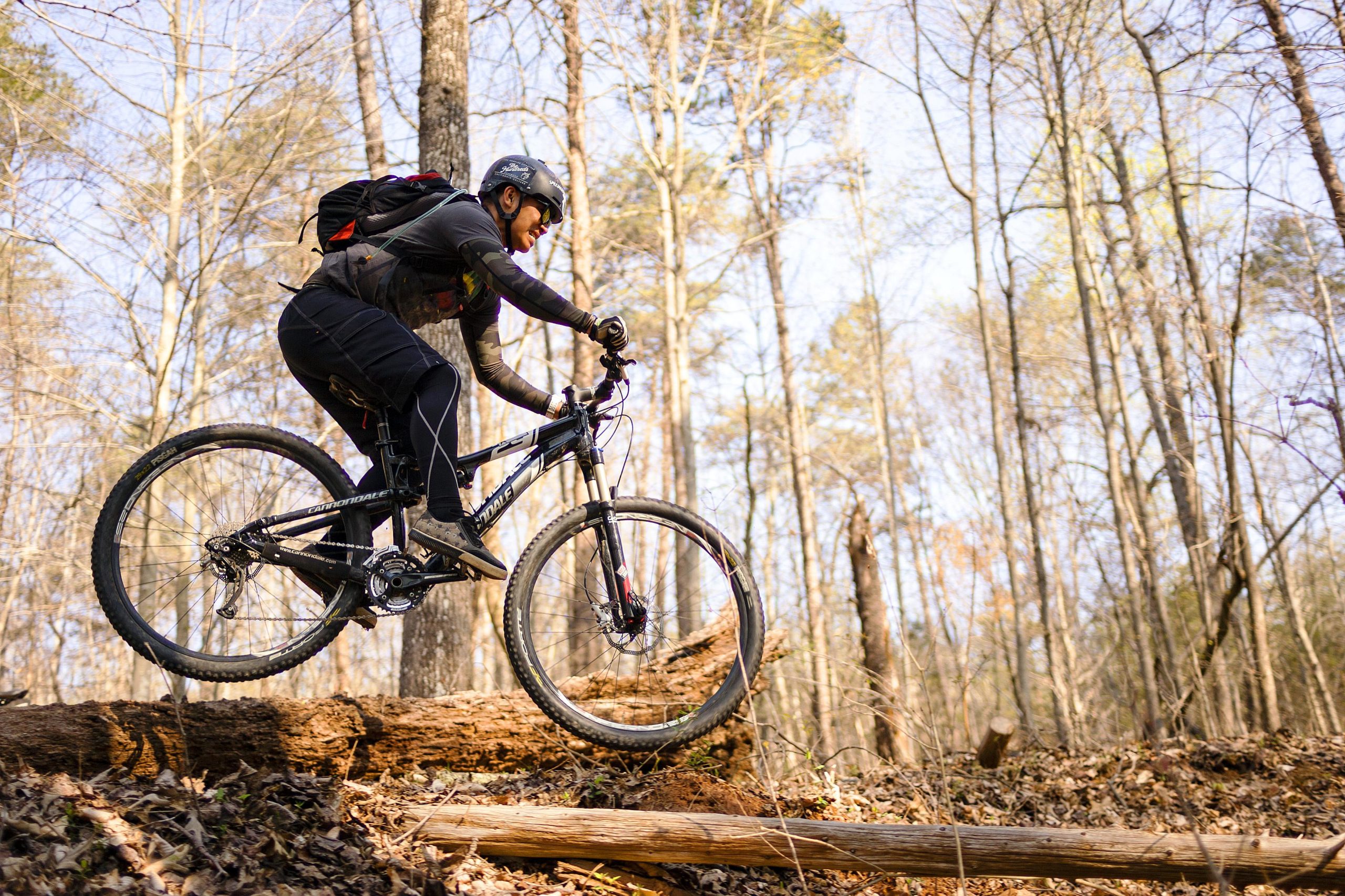 A mountain biker riding over a log in a wooded area during the day, surrounded by bare trees and a clear blue sky. The rider is wearing a helmet and riding gear, showcasing an adventurous moment in nature. Salem Lake mountain bike trail.