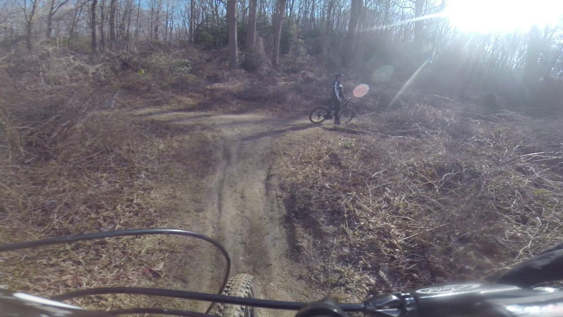 A view from a mountain biker's perspective on a dirt trail surrounded by trees, with a person on a bike in the background. The sun is shining brightly, creating lens flares and illuminating the natural landscape, which includes bare branches and dried foliage. Hartshorne Woods Park mountain bike trail.