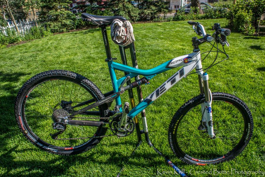 Yeti 575: A mountain bike with a blue and white frame is parked on a lawn. The bike features large tires, front suspension, and a visible gear mechanism. A tool and cloth are resting near the bike, indicating maintenance. The grassy background has trees and a fence in the distance.