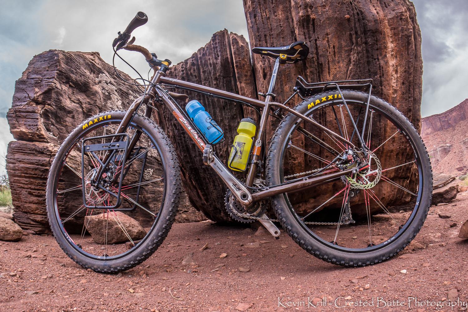 Salsa Fargo: A black mountain bike with yellow Maxxis tires, equipped with two water bottles (one blue, one yellow), is positioned against large rock formations in a rugged outdoor setting. The bike features a sturdy frame and a rear rack, surrounded by a natural landscape of reddish soil and dramatic sky.