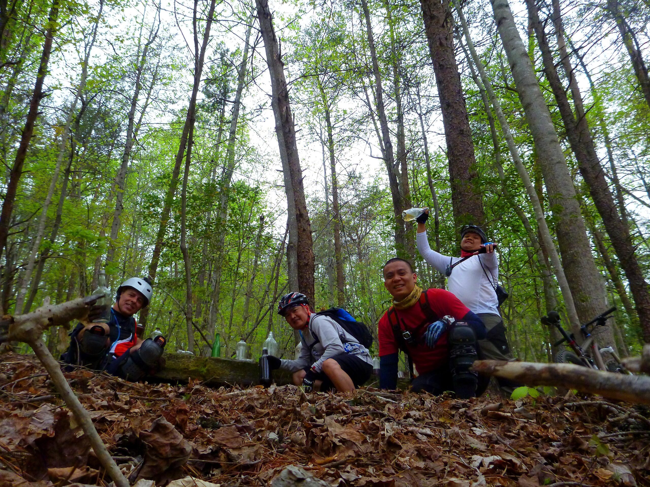 Four mountain bikers are taking a break in a wooded area. They are crouched on the forest floor, surrounded by green trees and fallen leaves. Two of them are smiling at the camera, while one holds up a bag, and the other appears to be adjusting his gear. Mountain bikes are visible in the background. Warrior Creek mountain bike trail.
