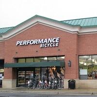 Photo of a brick building with a green roof, featuring the sign "Performance Bicycle" prominently displayed. The storefront has large windows showing bicycles inside, and several bikes are parked outside in front of the entrance.