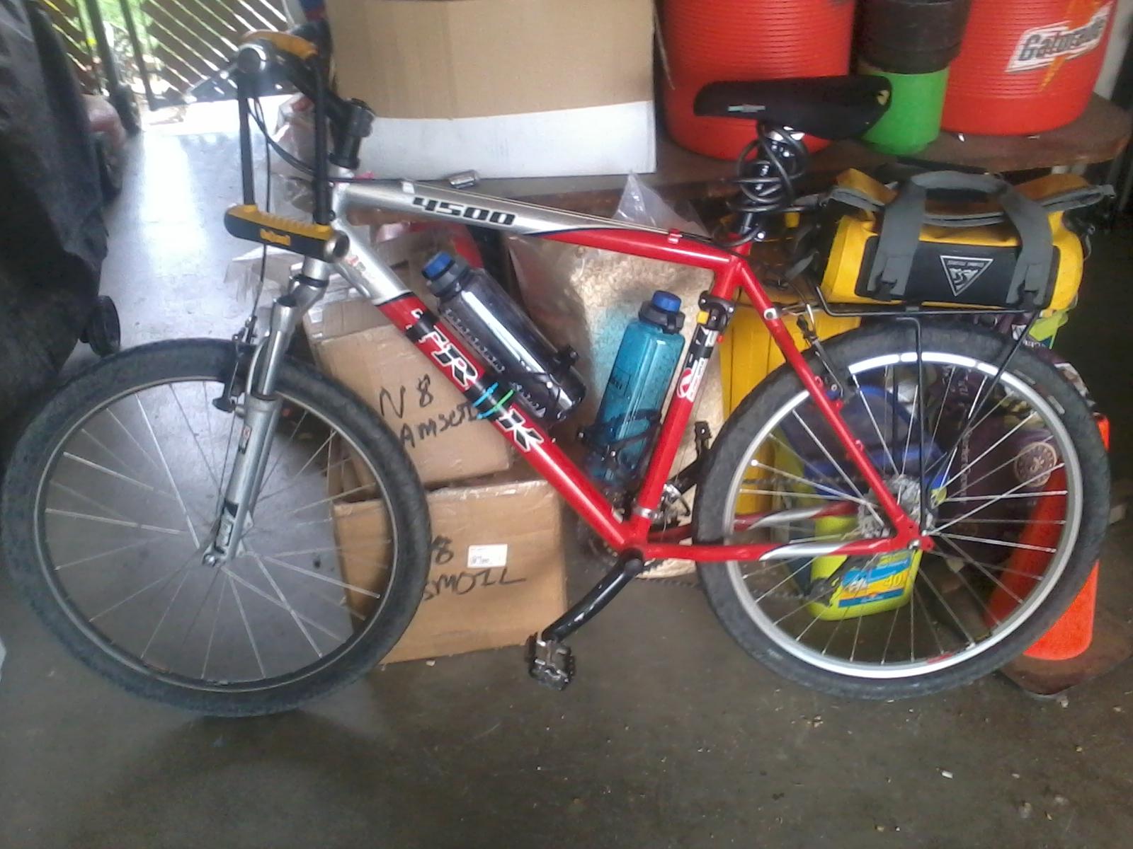Trek 4500: A red and silver mountain bike parked beside stacks of cardboard boxes in a garage. The bike features a yellow water bottle holder, two blue water bottles attached, and a black and yellow bag mounted on the rear rack. The background includes various storage items and containers, with soft lighting illuminating the scene.
