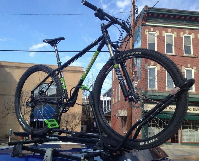 Salsa El Mariachi 3: A black mountain bike with green accents is secured on the roof rack of a vehicle. The bike features thick tires, suspension forks, and flat pedals. In the background, there are brick buildings and a clear blue sky.