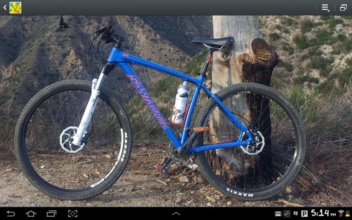 Santa Cruz Highball C: A blue mountain bike with a white front fork and large, treaded tires, leaning against a tree trunk with a scenic mountain background. A water bottle is secured in the bike's frame.