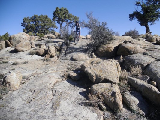 A person riding a mountain bike on a rocky terrain, with large boulders and sparse vegetation, under a clear blue sky.