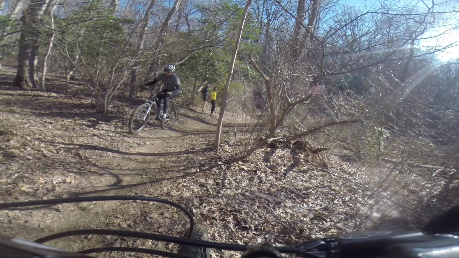 A mountain biker navigating a dirt trail through a wooded area, with a second person walking on the path in the background. The scene features a mix of bare trees and underbrush, with fallen leaves covering the trail, all under a clear blue sky. Hartshorne Woods Park mountain bike trail.